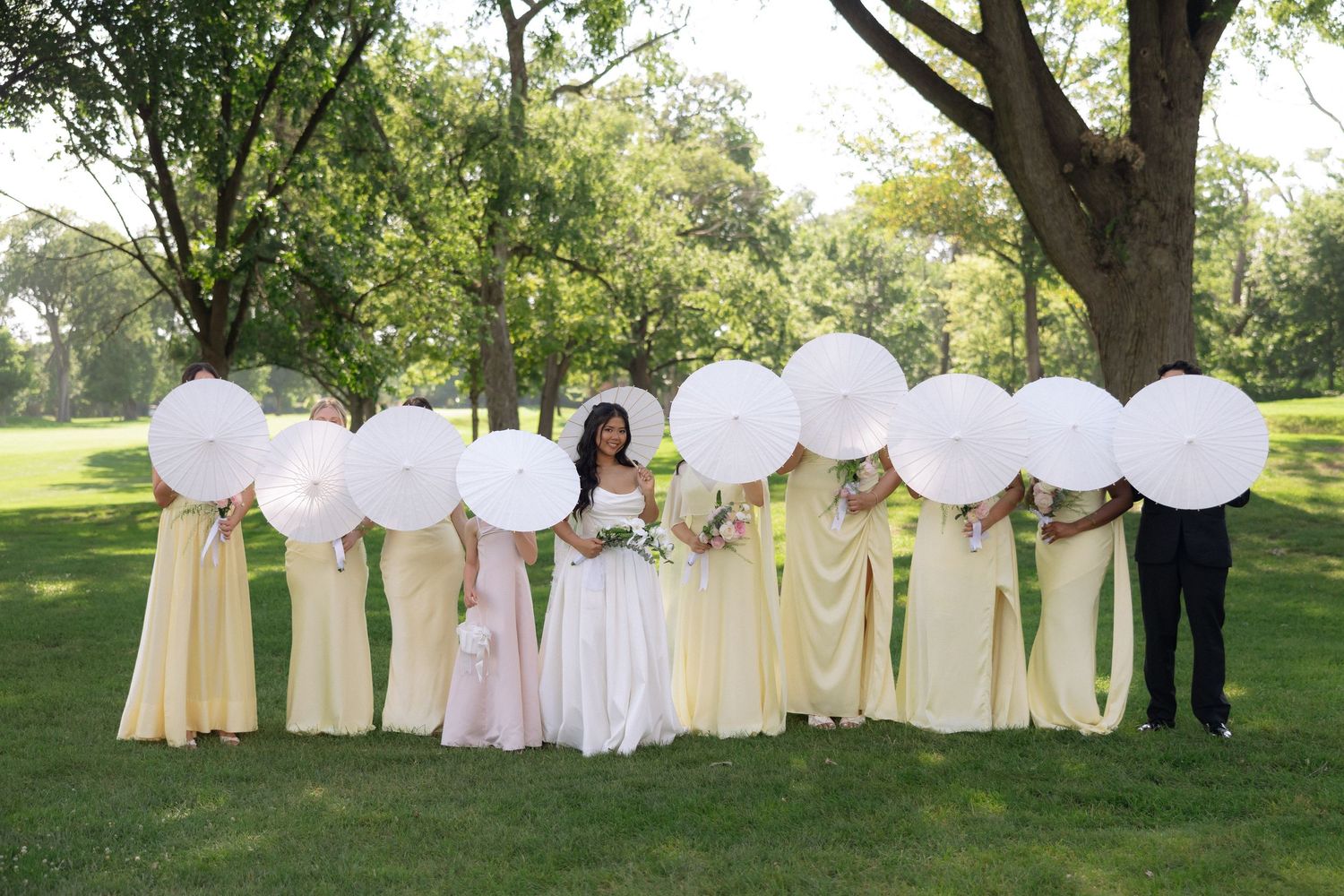 Bridesmaids in yellow dresses hiding faces behind white parasols with bride and groom outdoors.