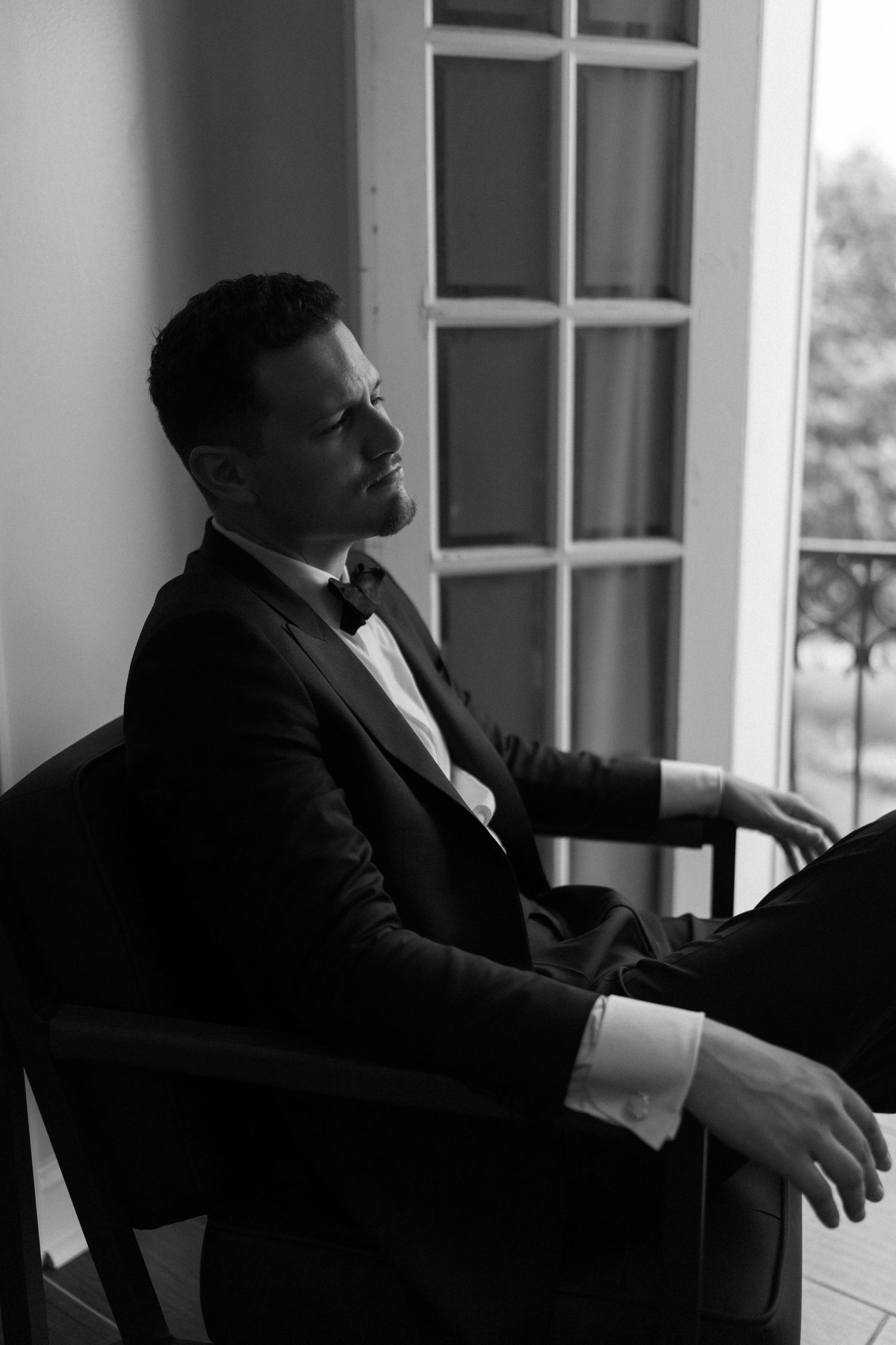 Black and white portrait of a well-dressed man in a tuxedo sitting in a chair near a window.