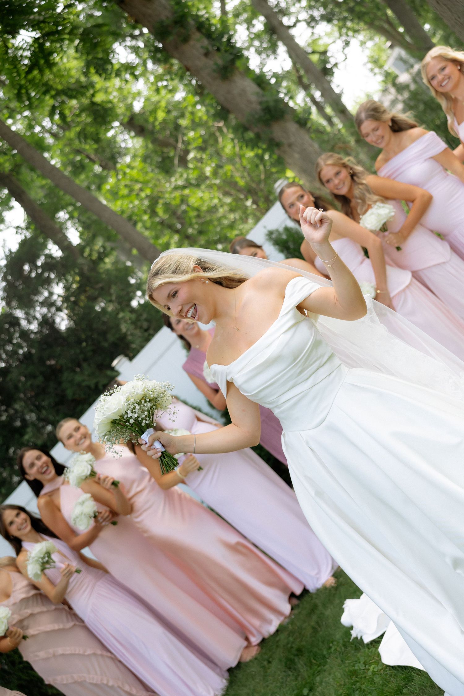 Laughing bride in white gown poses with bridesmaids in pink dresses holding white bouquets.