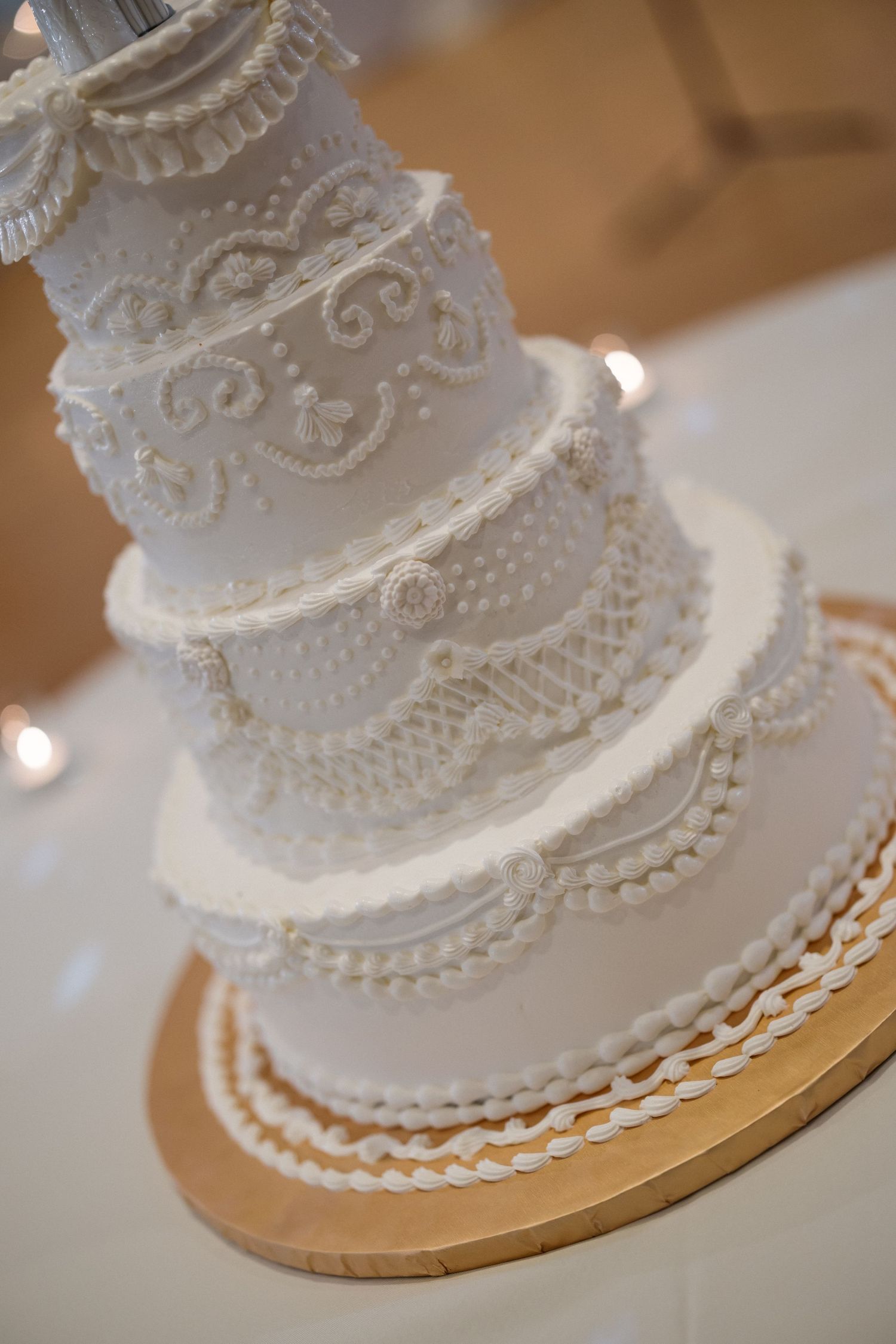 Elegant four-tier white wedding cake with intricate lace piping details on a gold cake board.