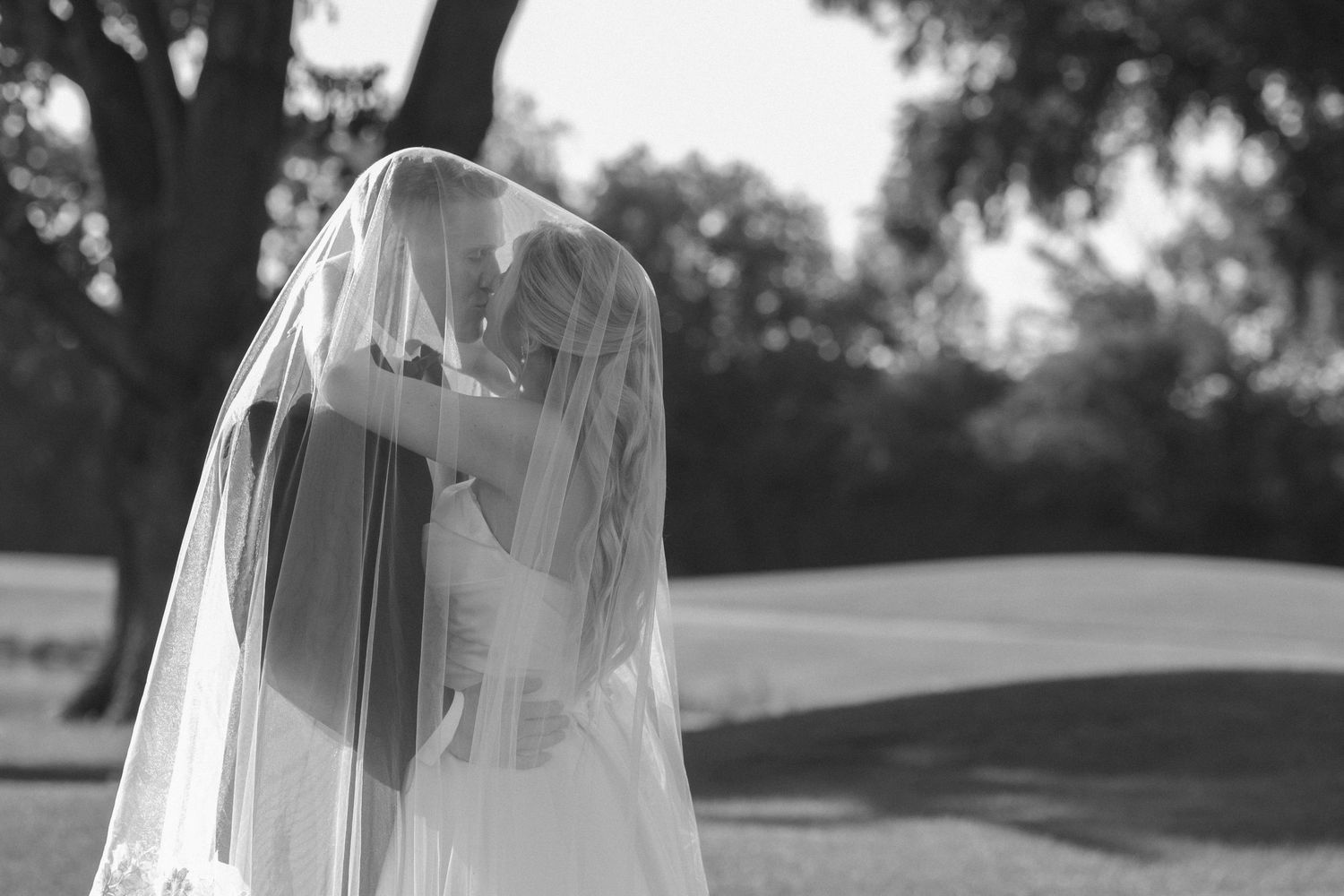 Black and white photo of bride and groom kissing under wedding veil outdoors among trees.