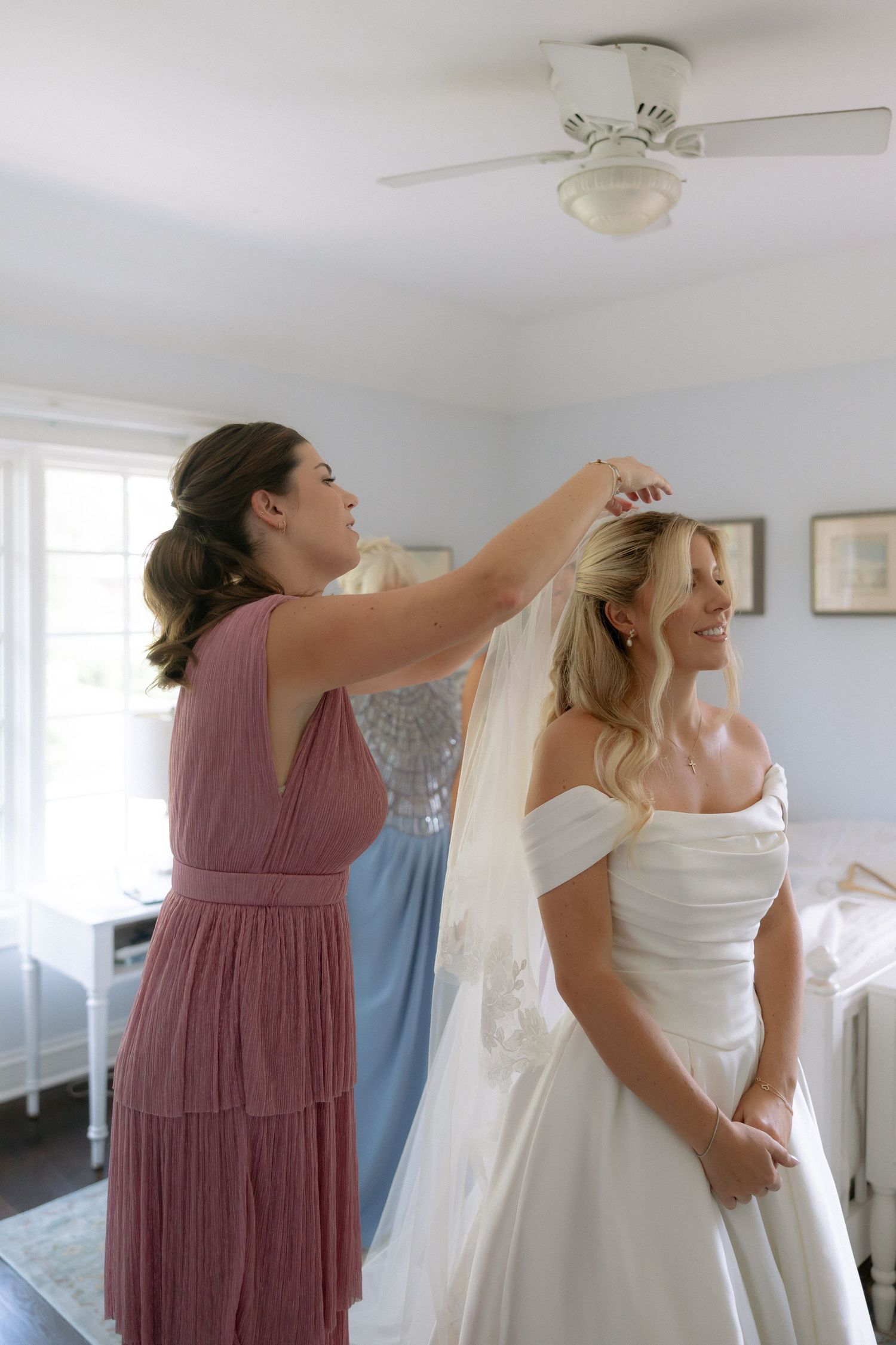 Bridesmaid in mauve dress adjusting the veil of a smiling bride in an off-shoulder white gown.