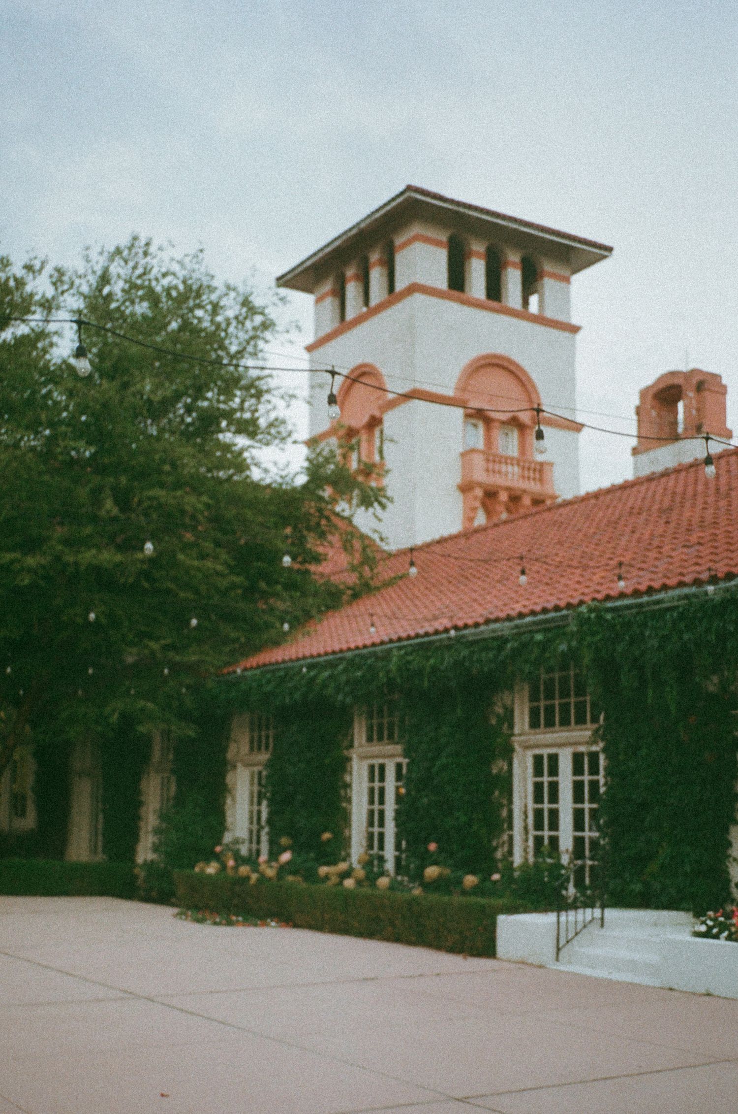 Historic building with red tile roof, ivy-covered walls, arched windows, and a white tower against a cloudy sky.