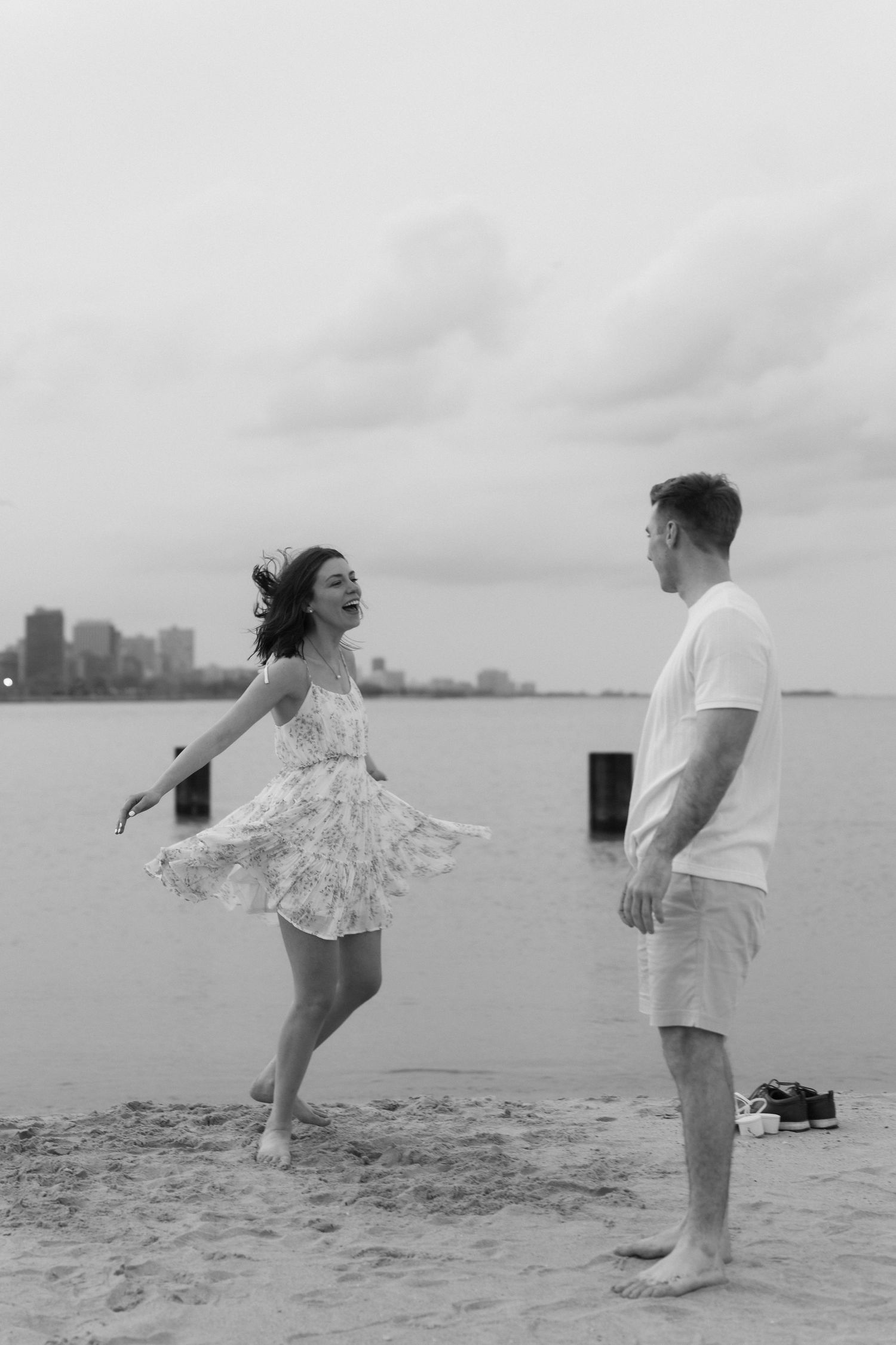 Black and white photo of a joyful couple dancing barefoot on a sandy beach with city skyline behind.