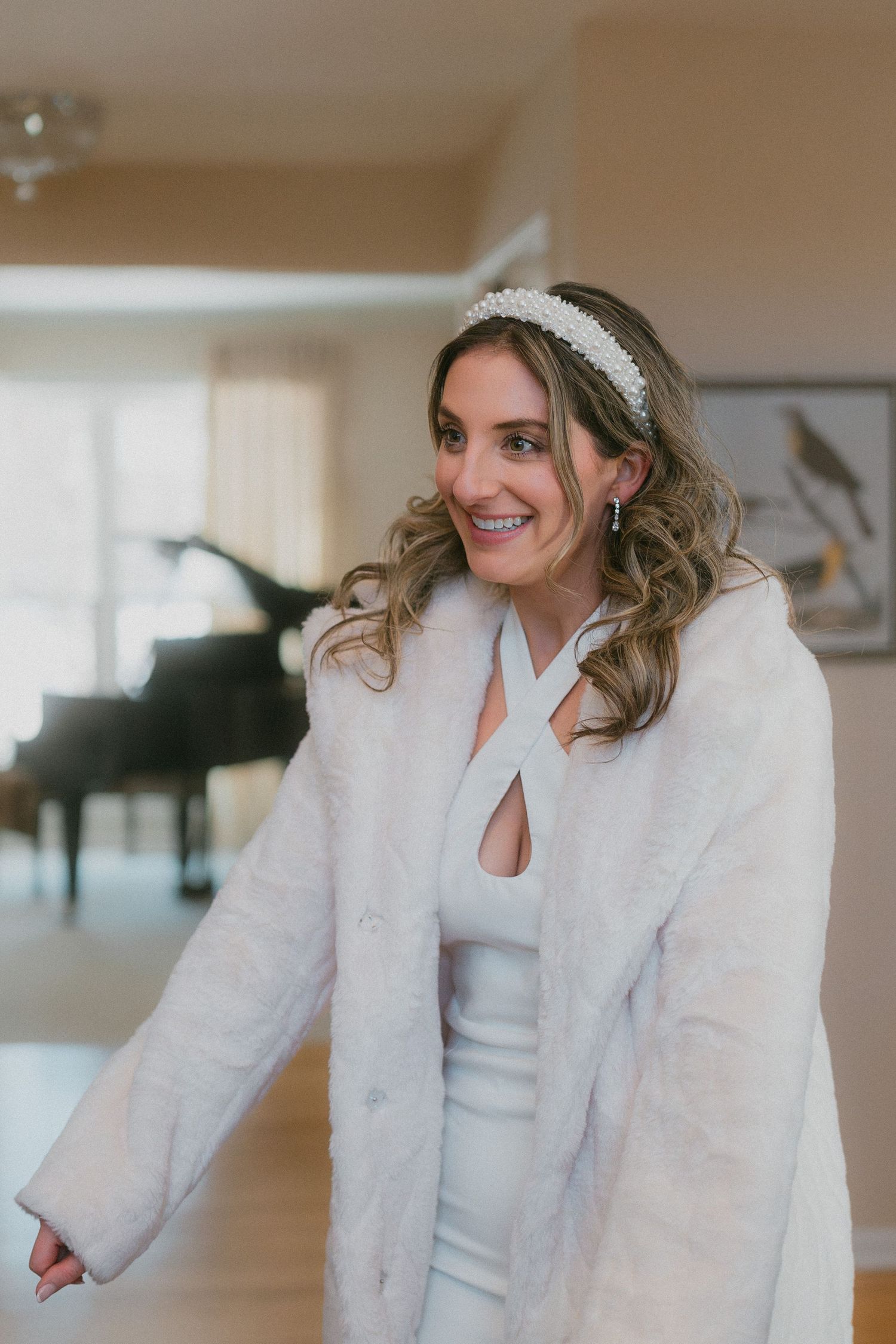Smiling bride wearing pearl headband and white fur coat stands in elegant home interior.