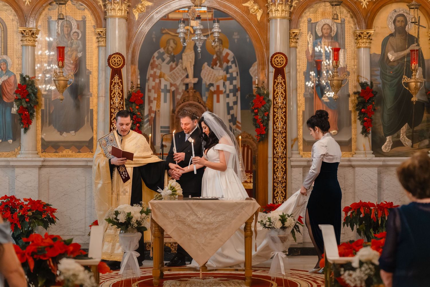 Bride and groom stand at altar during Orthodox wedding ceremony with priest reading scripture in decorated church.