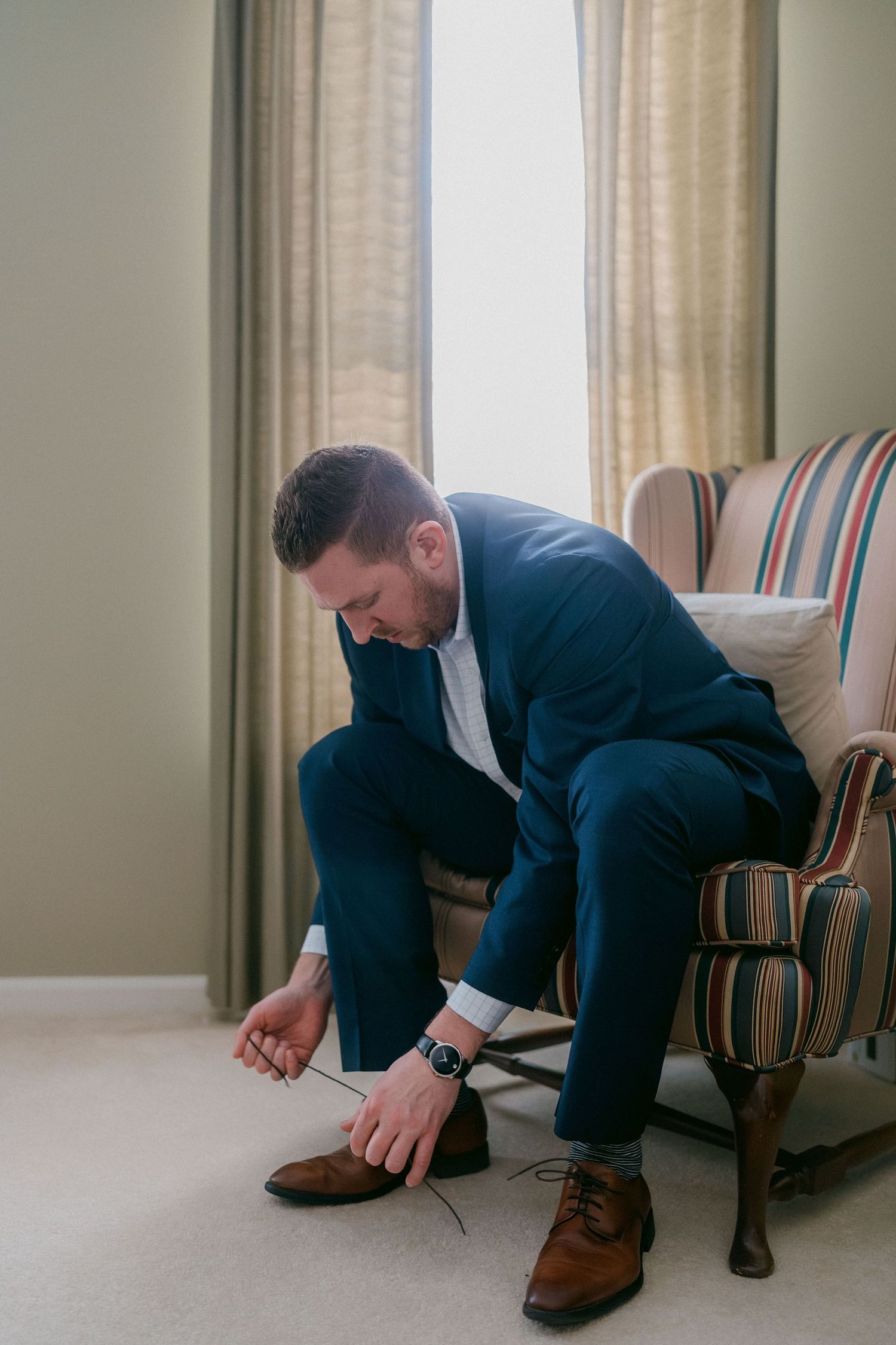 Groom in navy blue suit tying brown oxford shoes while seated in a striped armchair by a window.