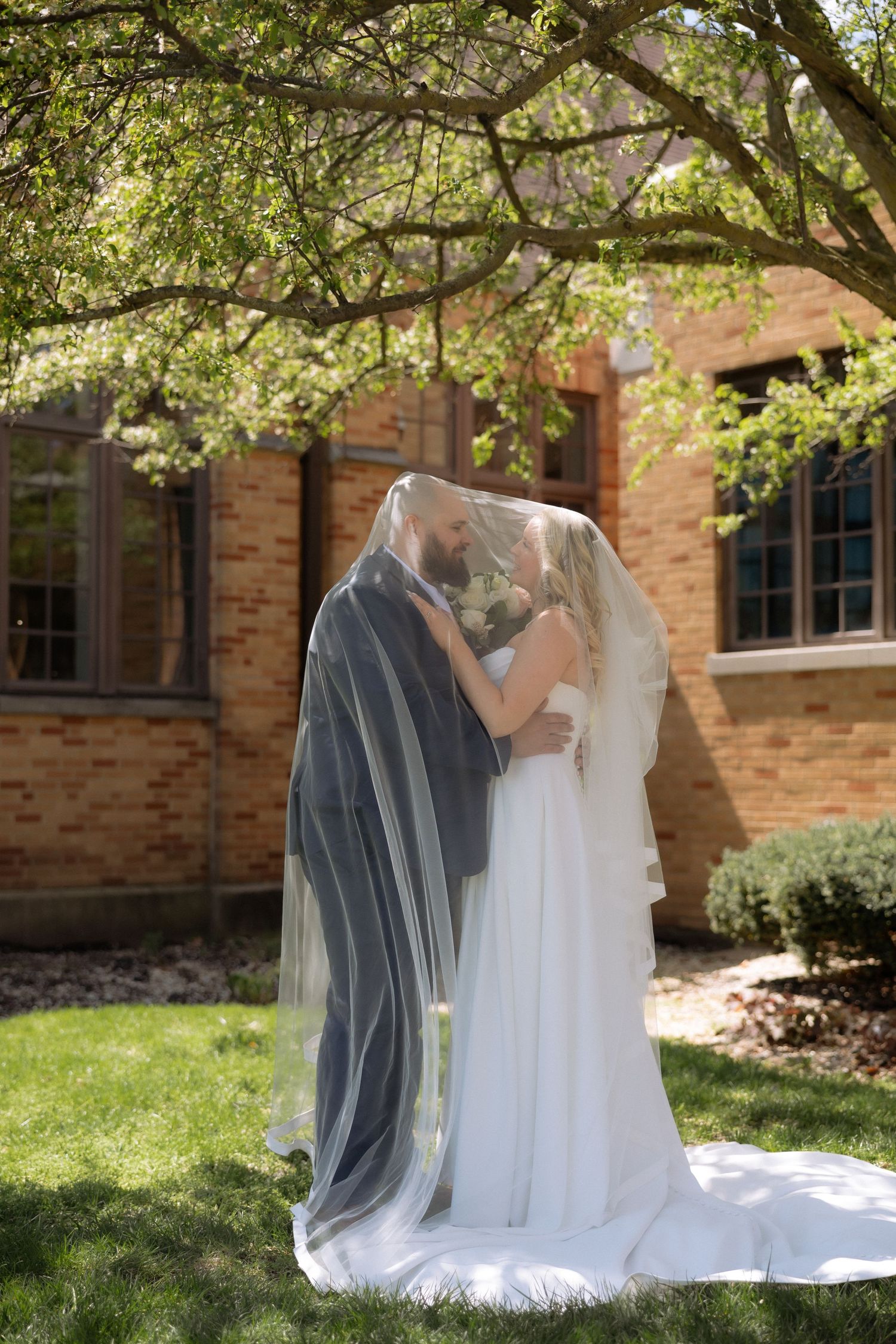 Bride and groom kissing under a flowing veil outdoors near a brick building surrounded by lush green trees.