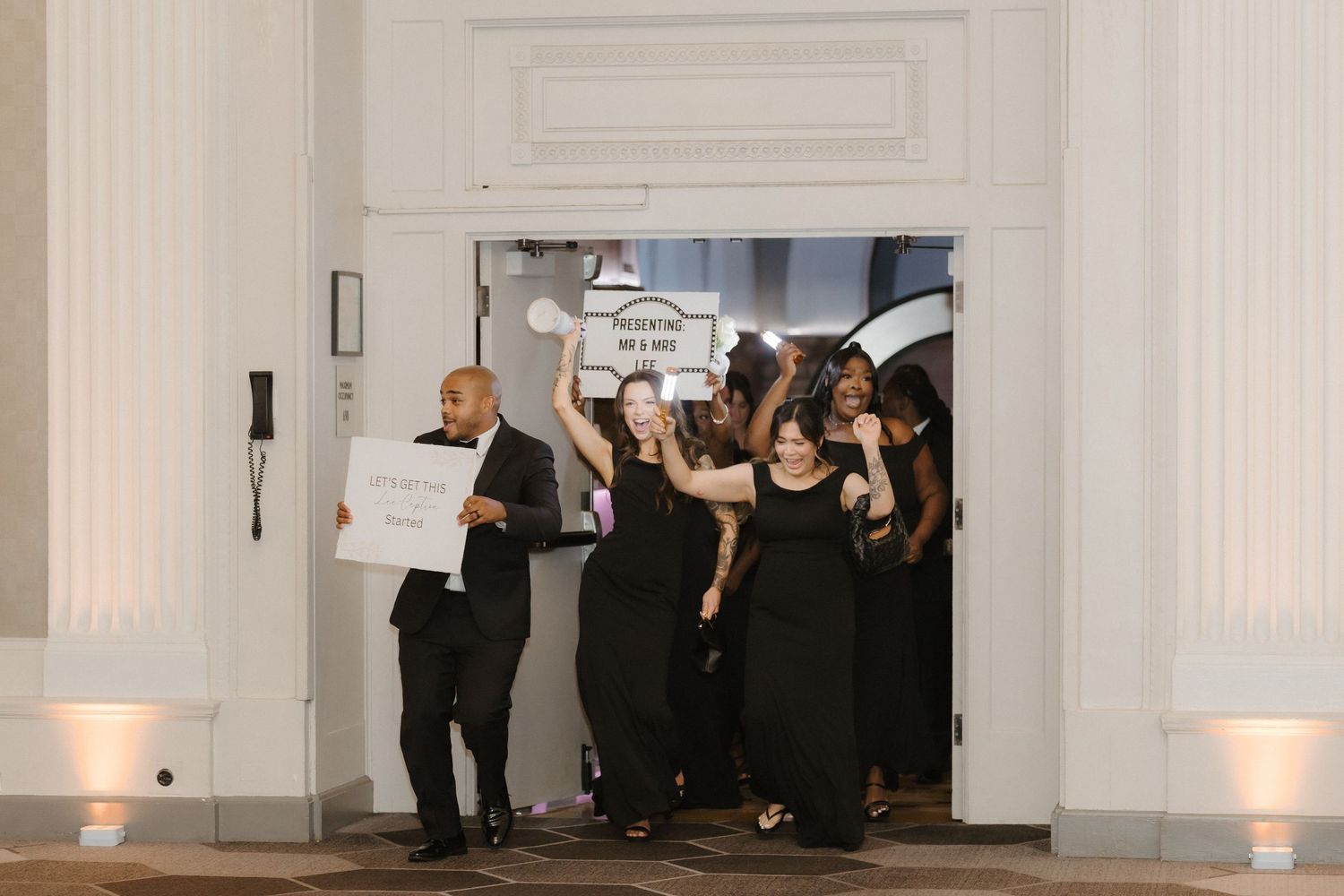 Wedding party in black attire exits elegant venue doors, holding signs and celebrating joyfully.