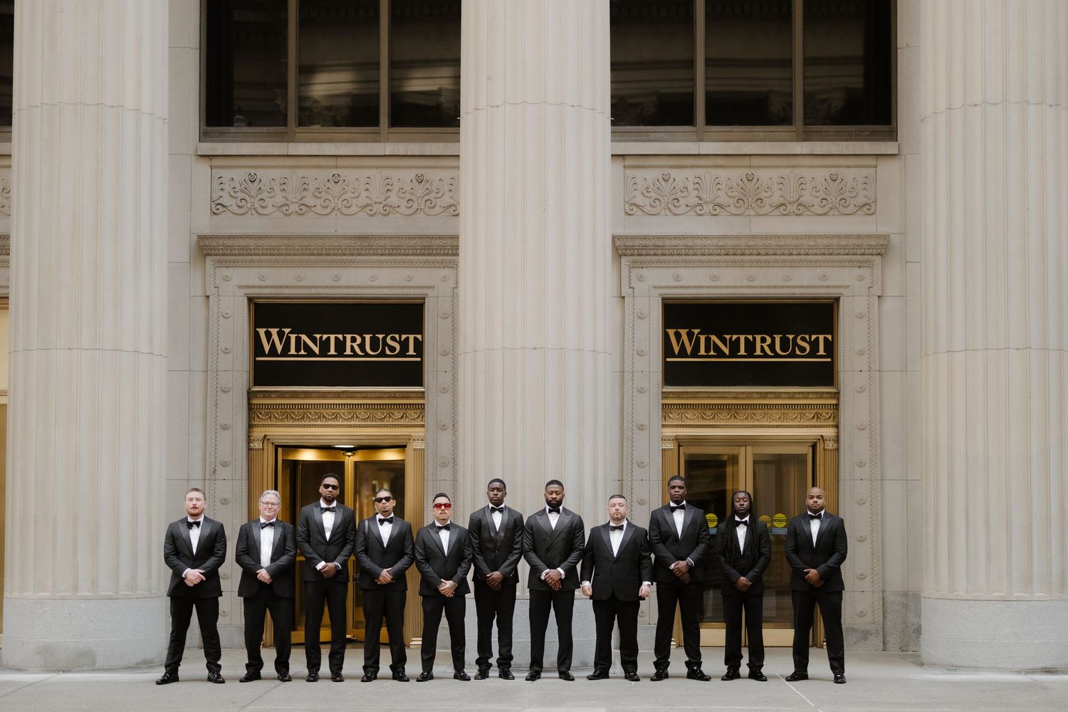 Groomsmen in black tuxedos posing in front of Wintrust building columns for a wedding party photo.