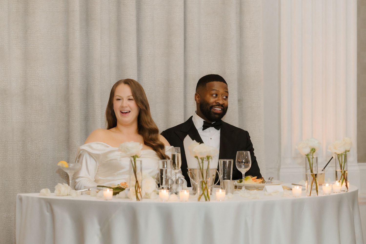 Bride and groom laughing at sweetheart table with white floral centerpieces and candles at elegant wedding reception.