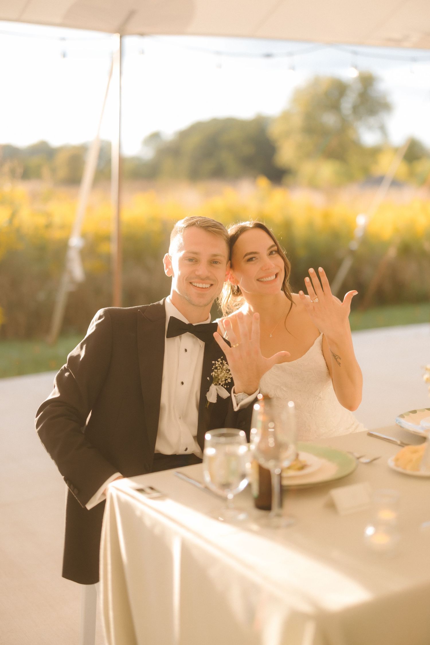 Happy newlyweds smile and show off wedding rings at their sweetheart table with golden wildflower fields behind them.
