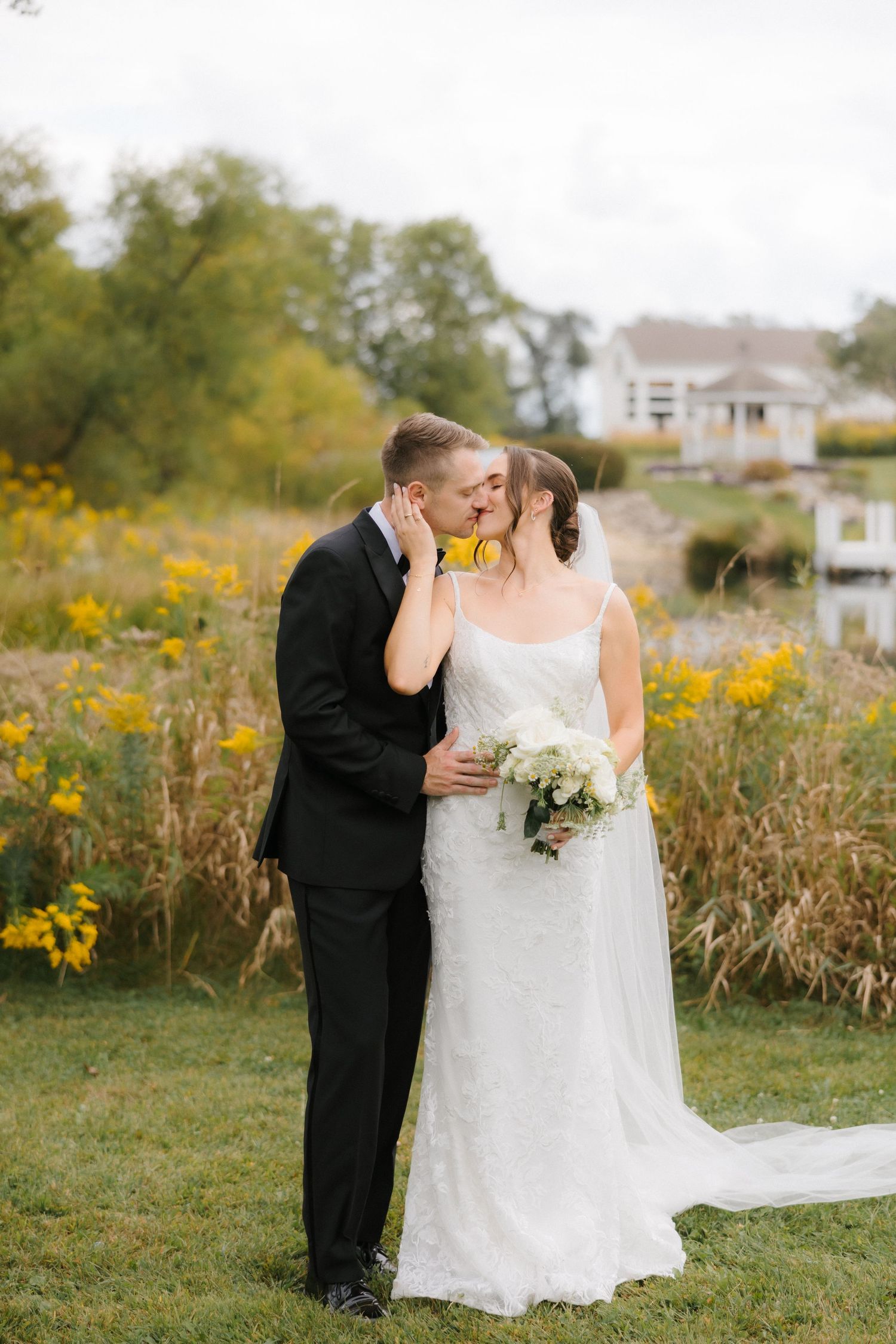 Bride and groom share a kiss outdoors surrounded by yellow wildflowers, bride holding white bouquet in wedding gown.