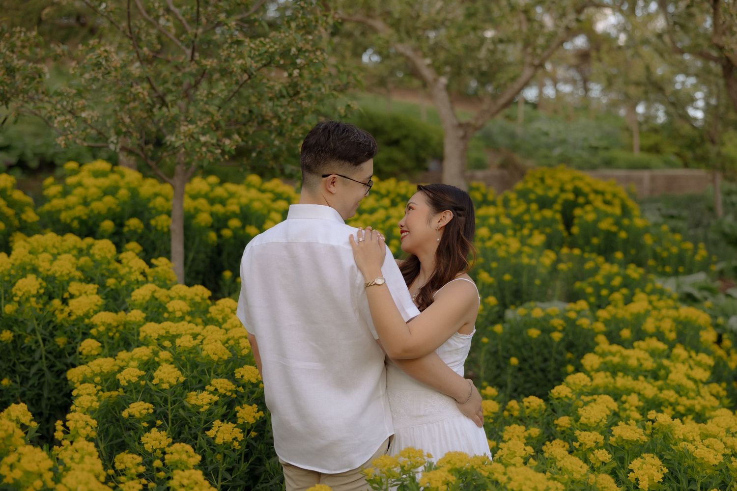 Couple embracing romantically among vibrant yellow flowers in a lush garden, both dressed in white outfits.