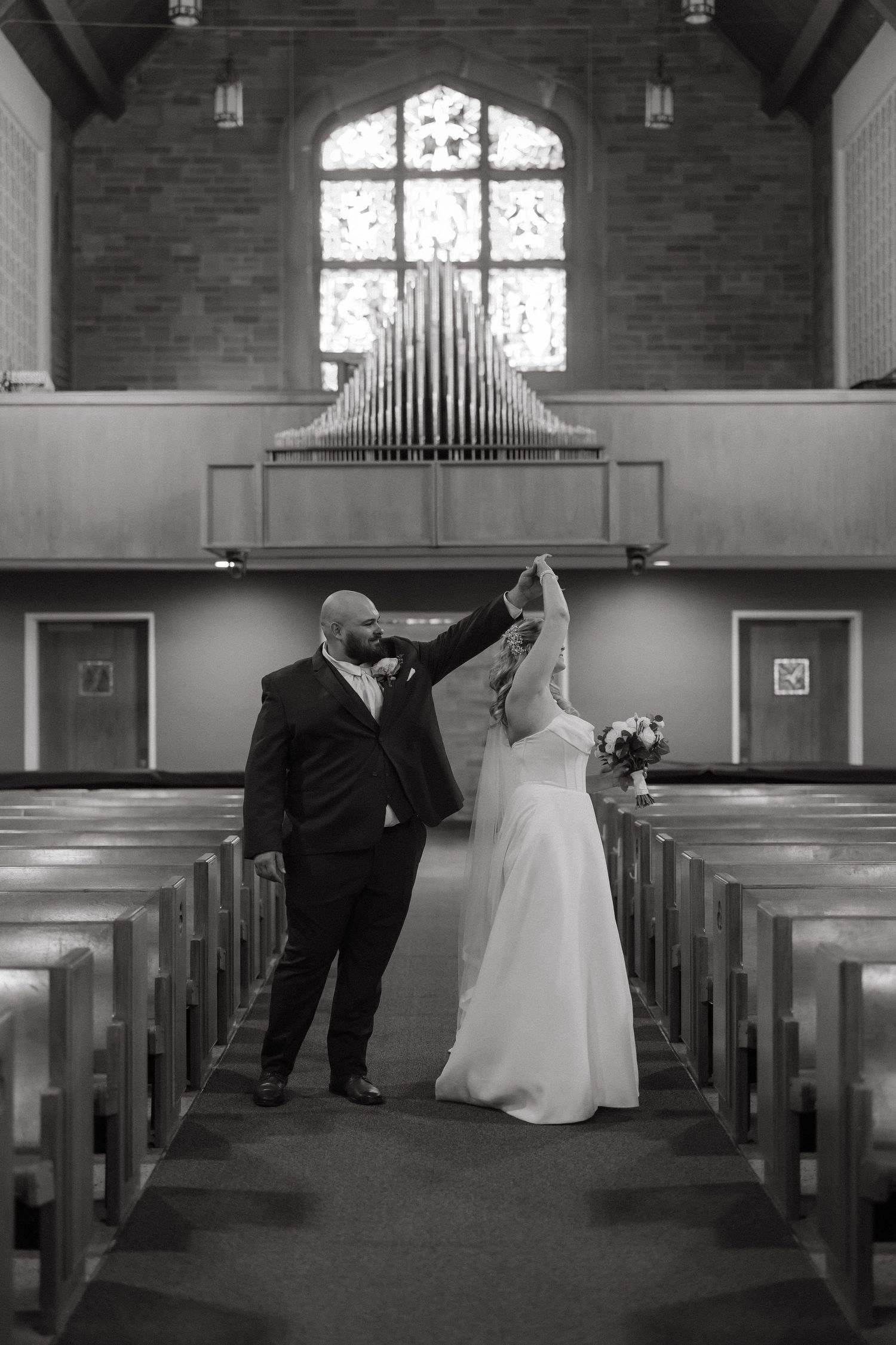 Black and white photo of a bride and groom dancing in a church aisle, with organ pipes and stained glass window behind them.