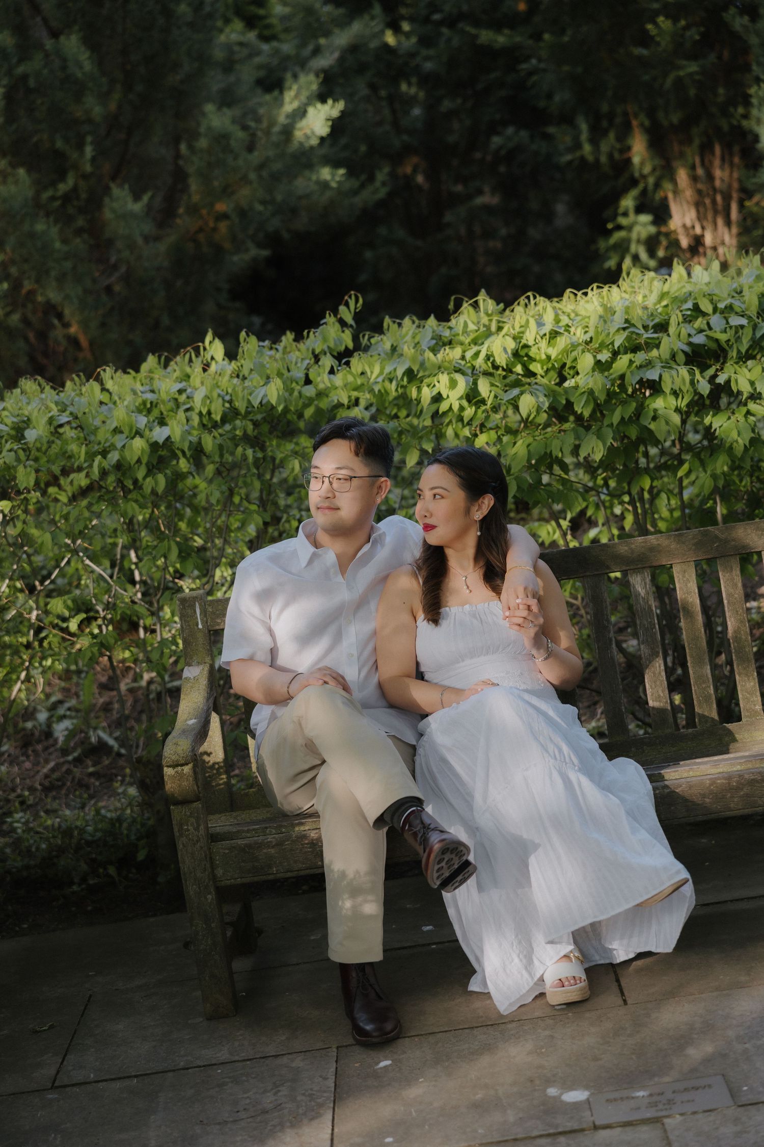 A couple sits closely on a wooden bench in a lush garden, wearing white outfits, sharing a romantic moment.