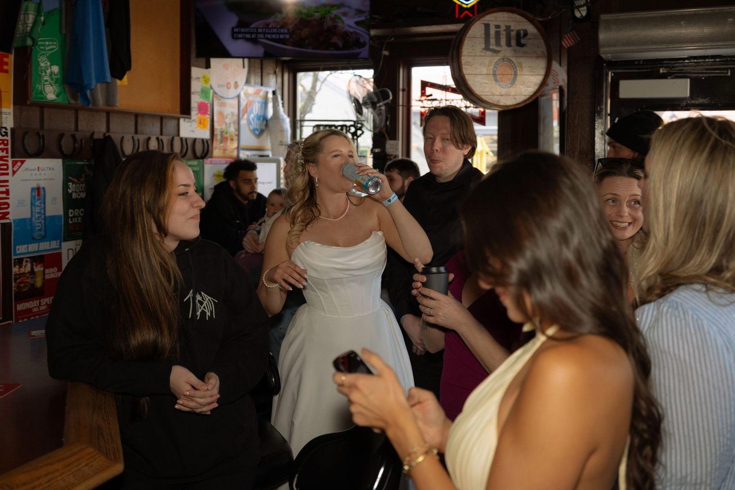 Bride in white dress drinking at a bar celebration with friends gathered around in a lively venue.