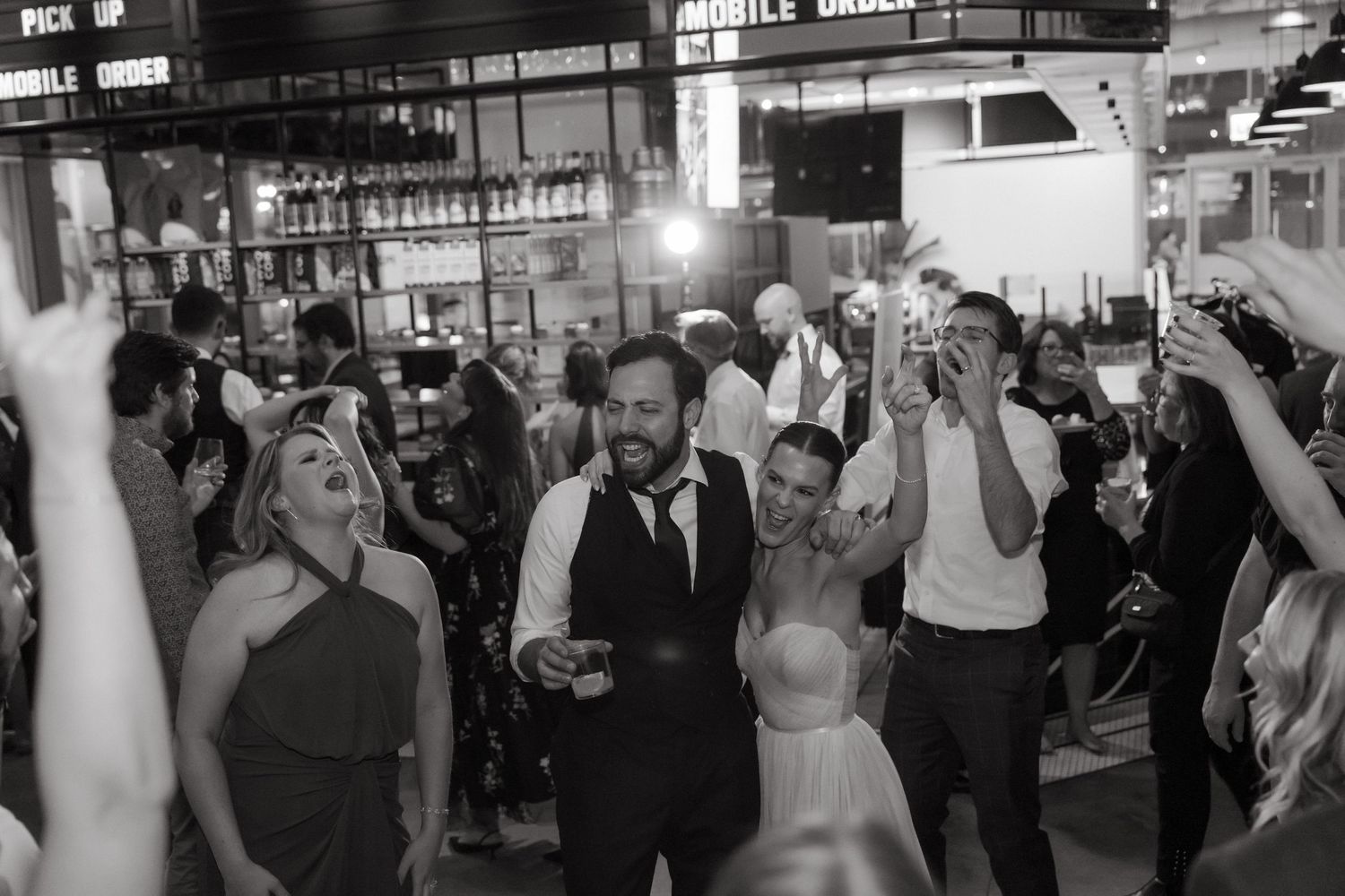 Black and white photo of bride and groom dancing with guests at lively wedding reception.