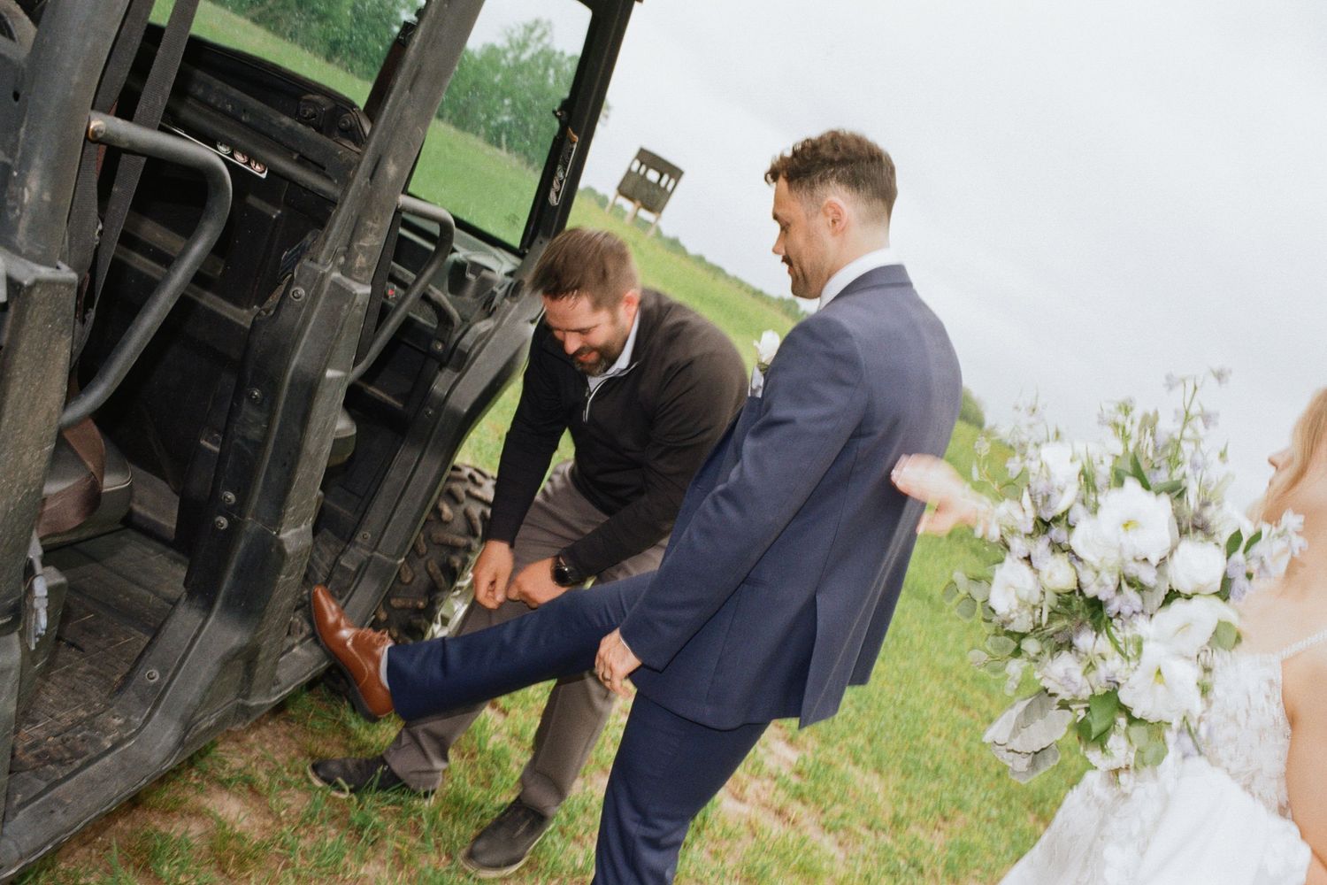 Groom in blue suit gets shoe fixed by man near military vehicle at outdoor wedding ceremony.