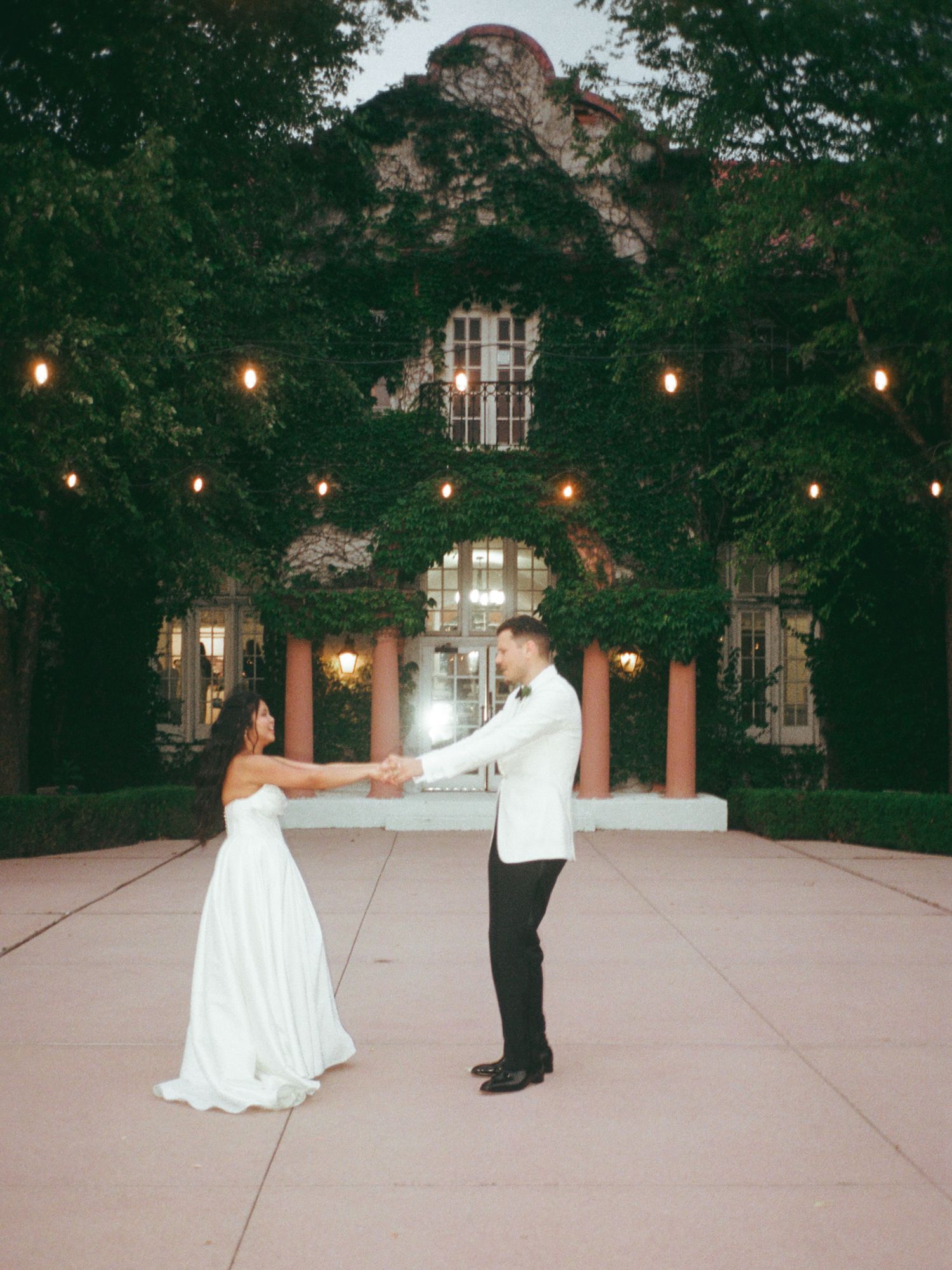 A bride and groom hold hands and dance outside an ivy-covered venue with string lights at dusk.