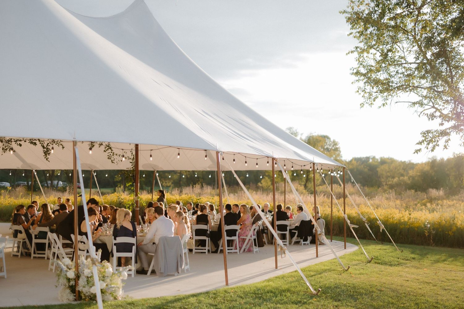Elegant outdoor wedding reception under a white sailcloth tent surrounded by lush greenery at golden hour.