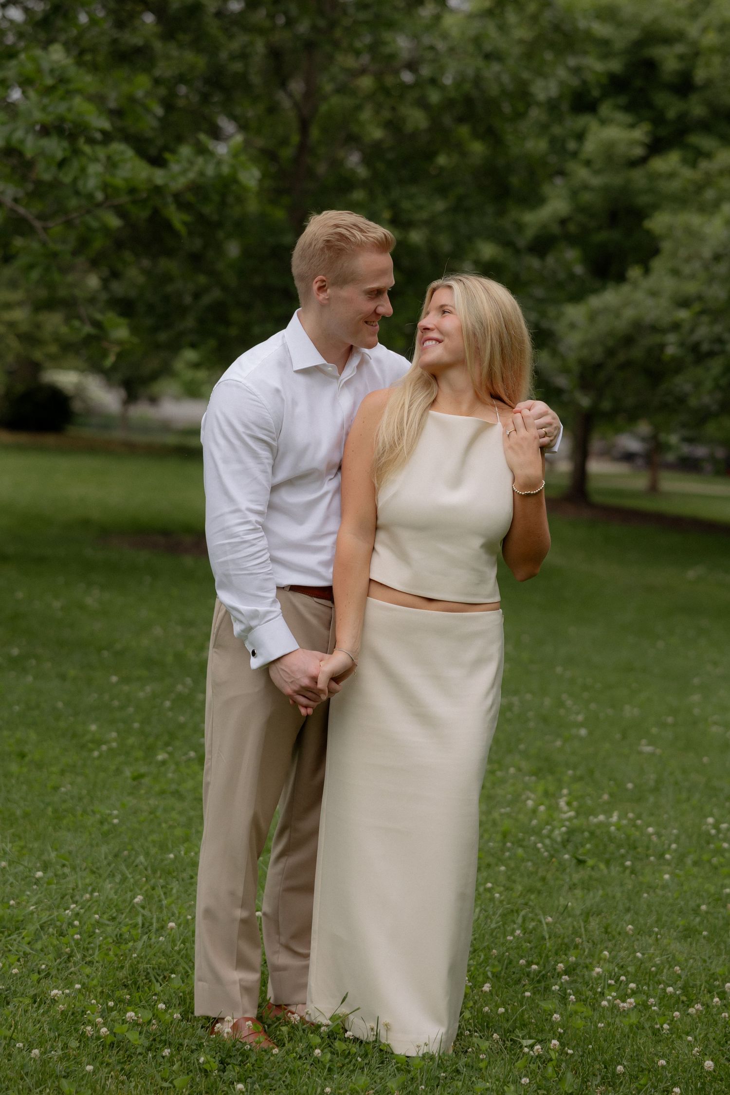 A smiling couple holds hands in a lush green park, the man in white shirt and the woman in a cream outfit.