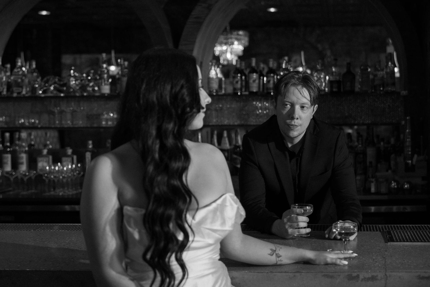 Black and white photo of a man in a suit at a bar conversing with a woman in a white dress.