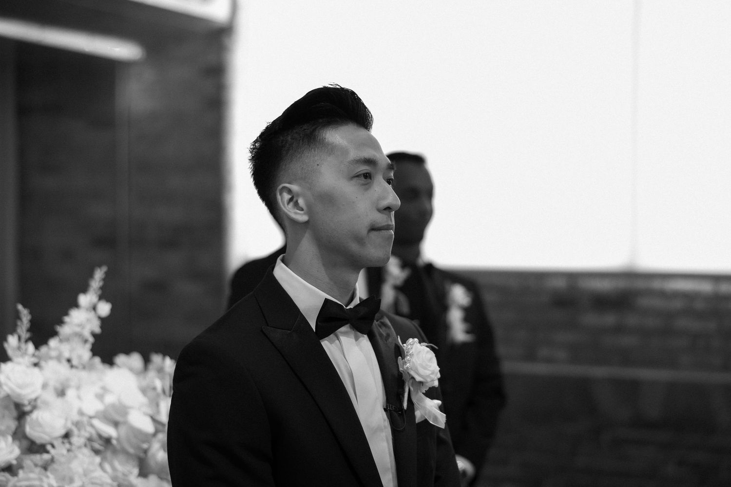 Groom in black tuxedo with boutonniere stands at wedding altar in black and white photo.