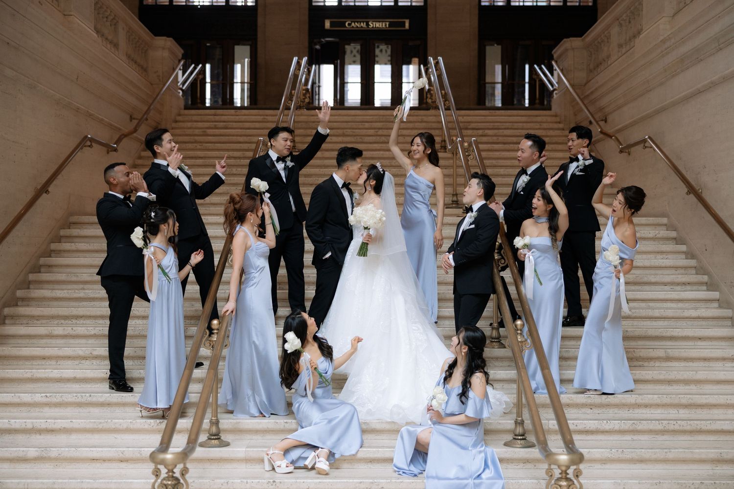 Wedding party celebrates on grand staircase with bridesmaids in blue gowns and groomsmen in tuxedos.