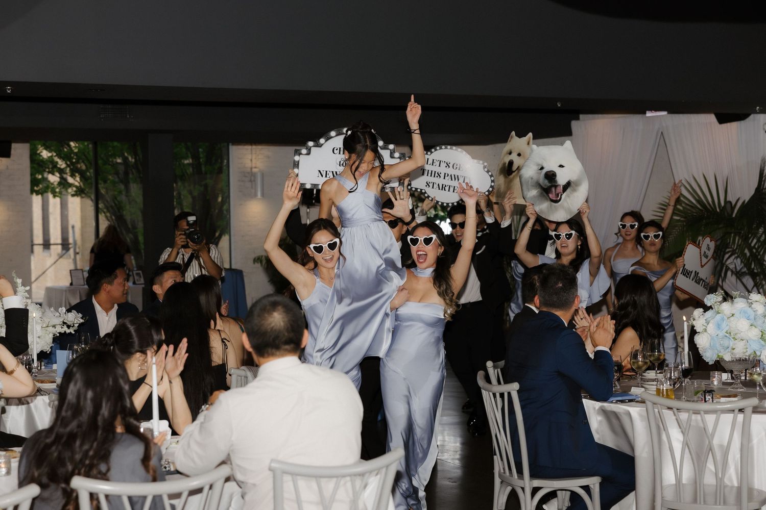 Bridesmaids in blue dresses and sunglasses dance energetically at a wedding reception with white floral centerpieces.