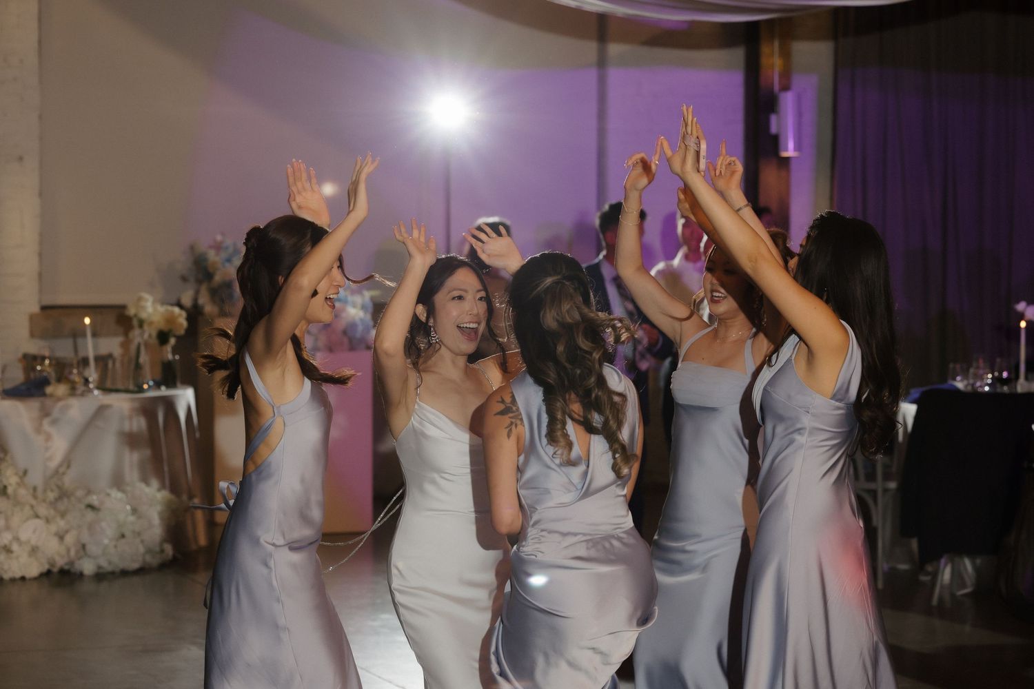 Bride and bridesmaids in silver gowns dancing joyfully together on the wedding reception dance floor.