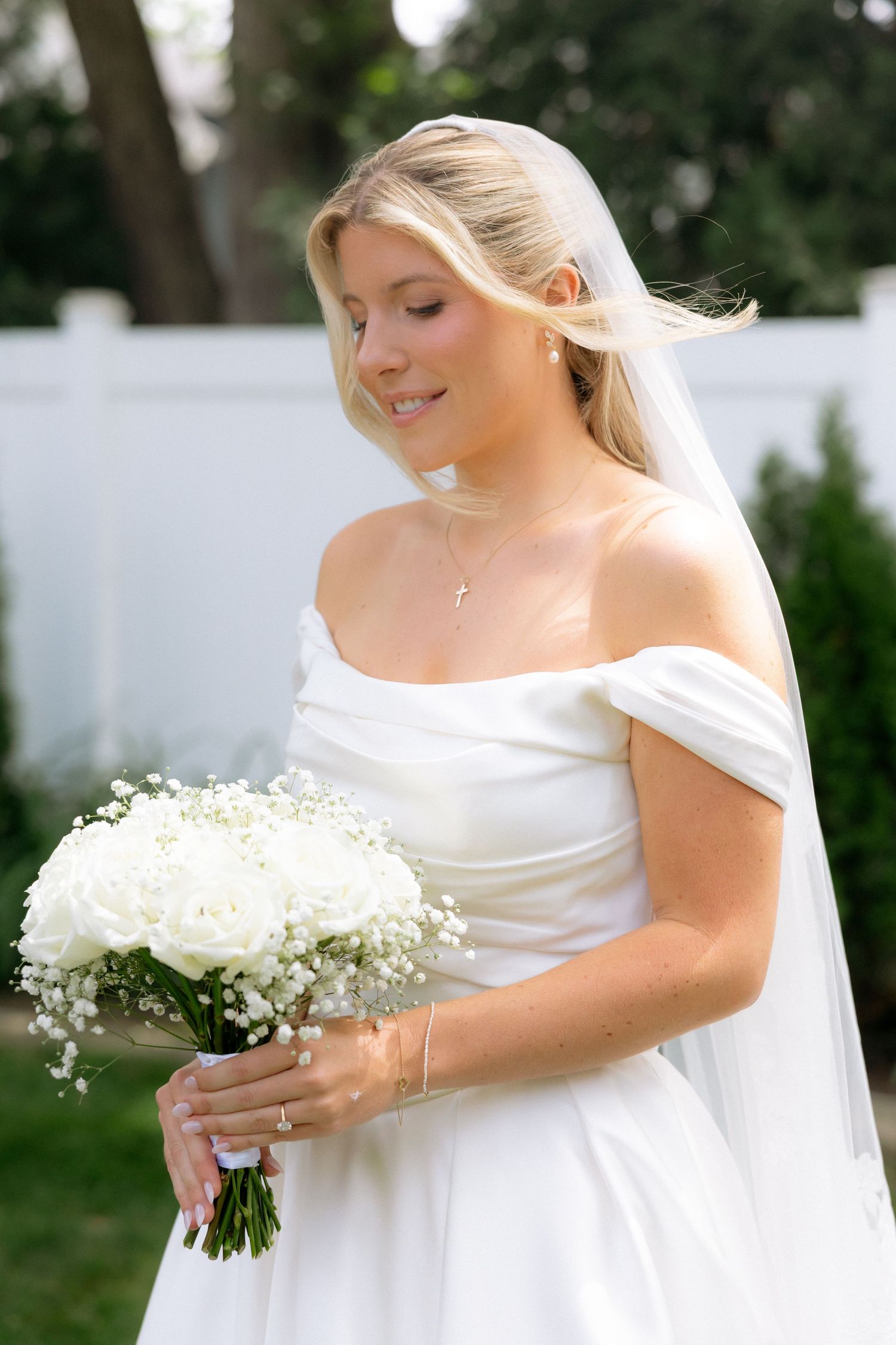 Blonde bride in off-shoulder white satin gown holds white rose and baby's breath bouquet outdoors.