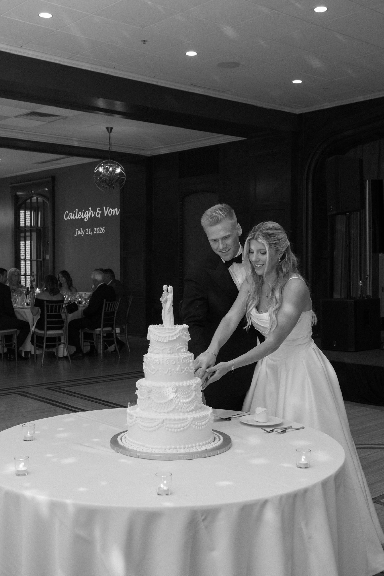 Bride and groom cut their elegant tiered wedding cake at reception, names projected on wall behind them.