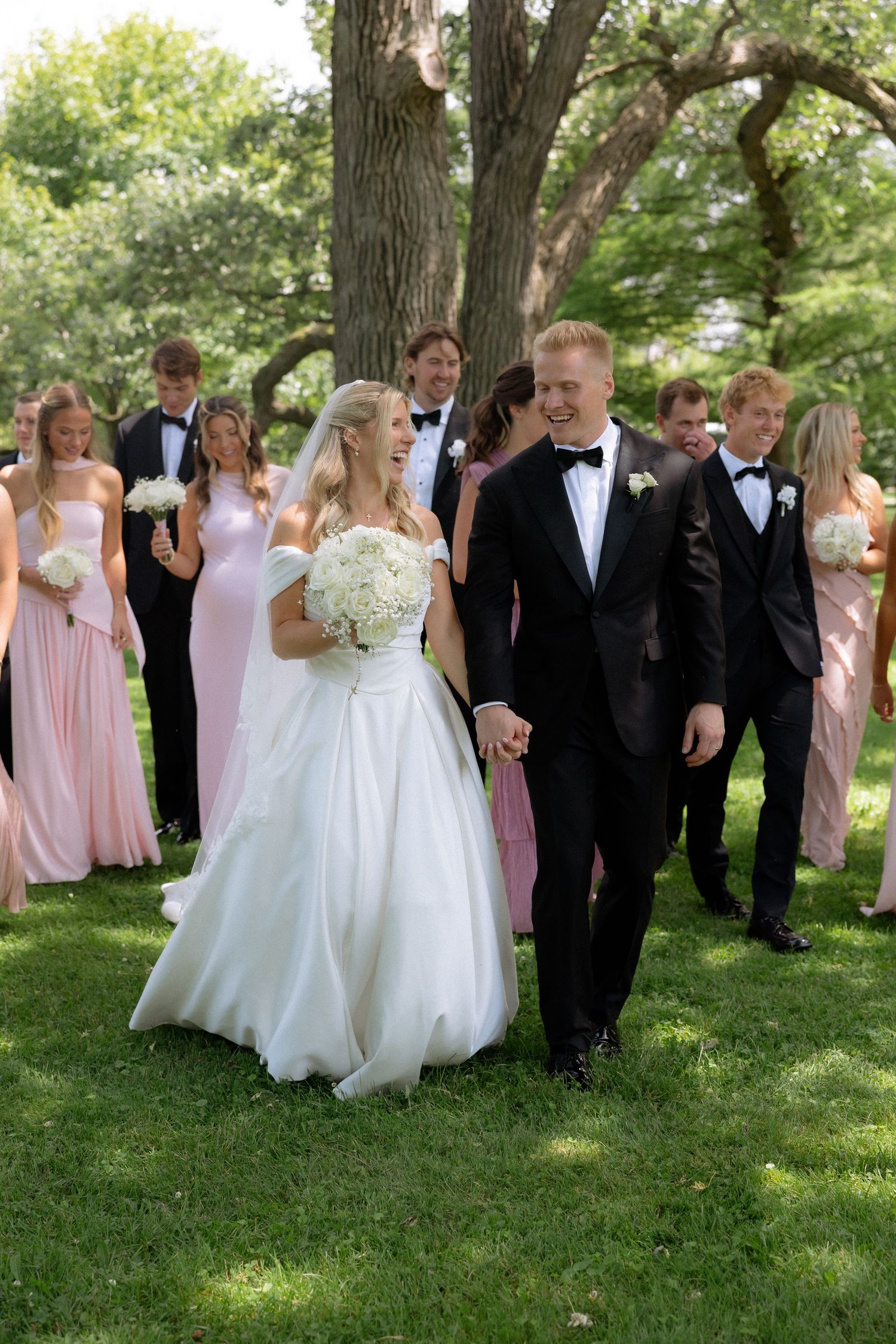 Wedding party walks outdoors, bride in white gown holds bouquet, groom in tuxedo, bridesmaids in pink dresses.
