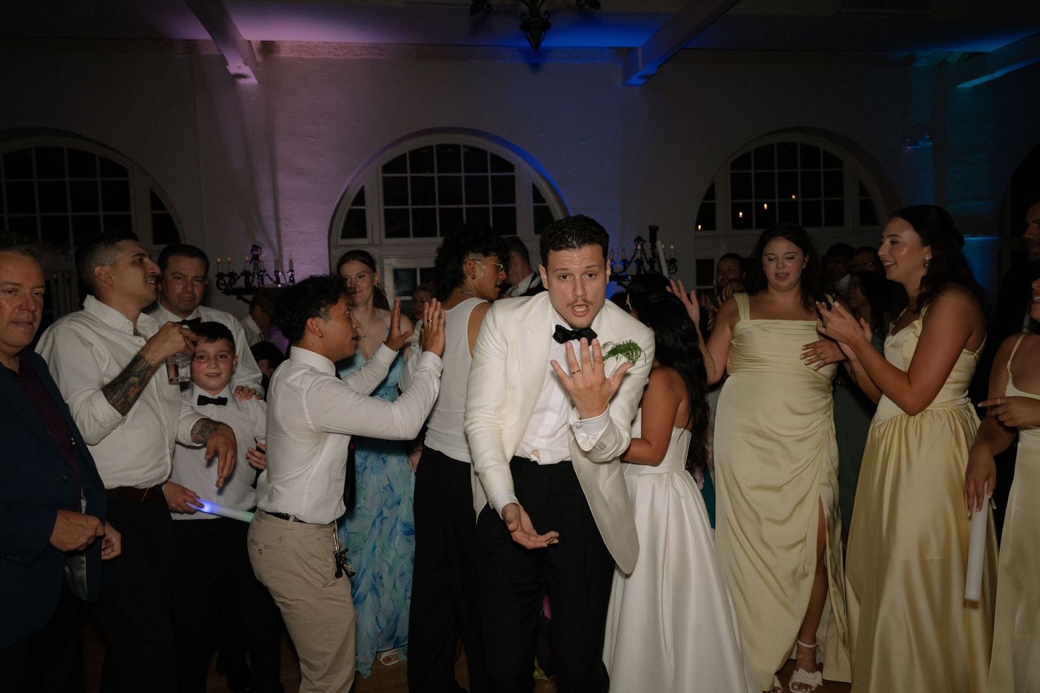 Groom dances joyfully surrounded by wedding guests on illuminated blue-lit reception dance floor.
