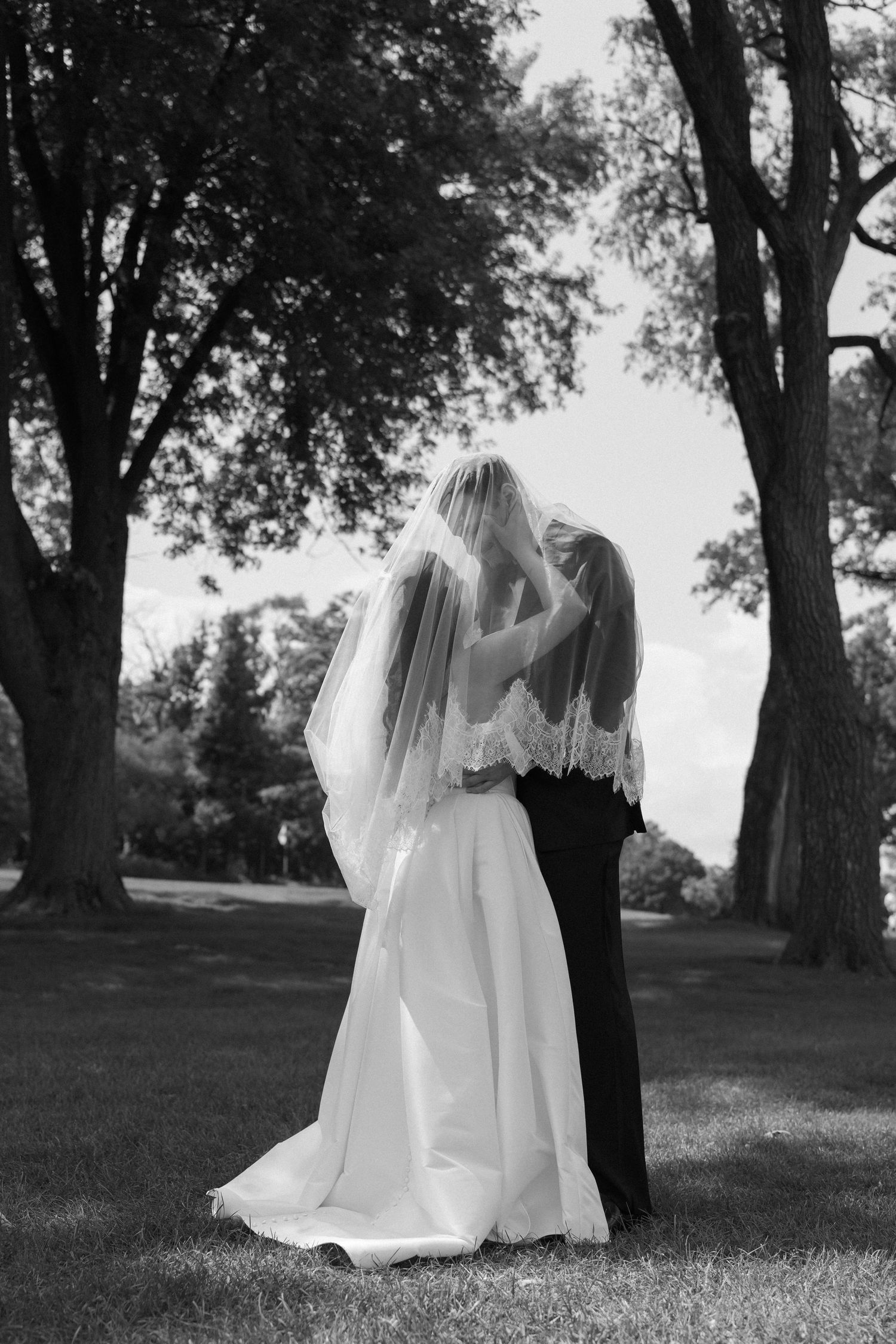 Bride and groom share romantic kiss under trees in black and white wedding portrait outdoors.
