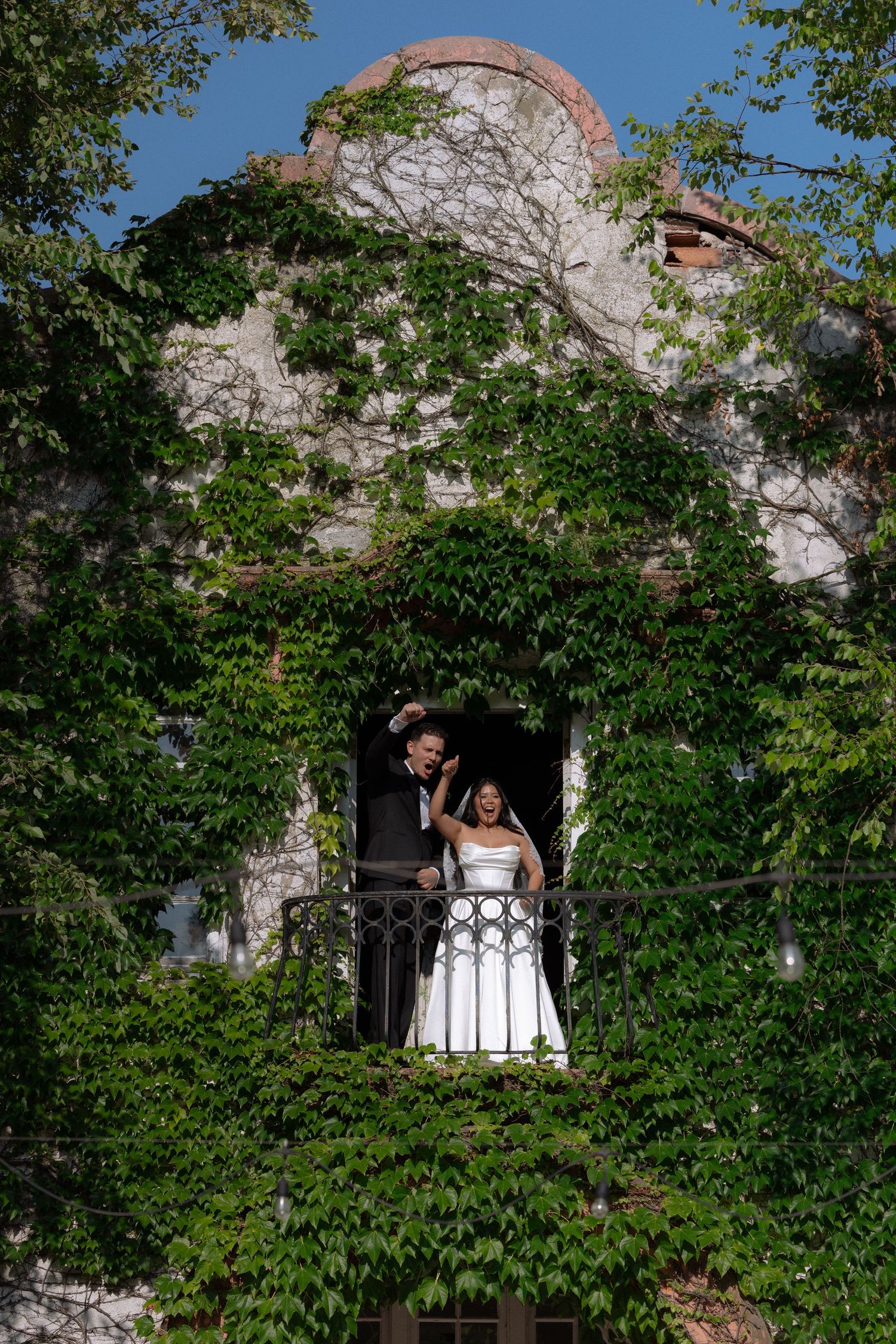 Joyful bride and groom pose on an iron balcony of an ivy-covered stone building on their wedding day.