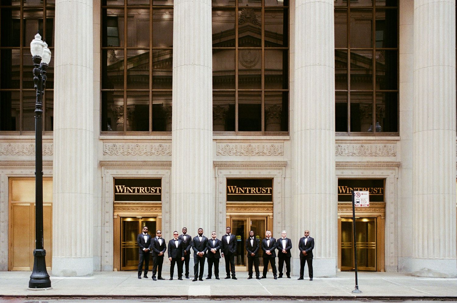 Groomsmen in black tuxedos posing in front of Wintrust building with grand columns in Chicago.