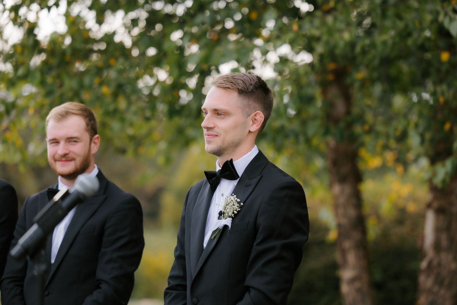Groom in black tuxedo smiles warmly during outdoor wedding ceremony with lush green trees in background.