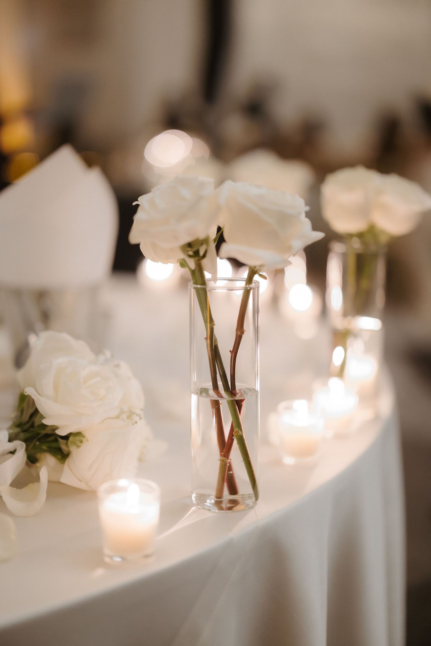 White roses in glass vases with lit candles on a white tablecloth at an elegant wedding reception.