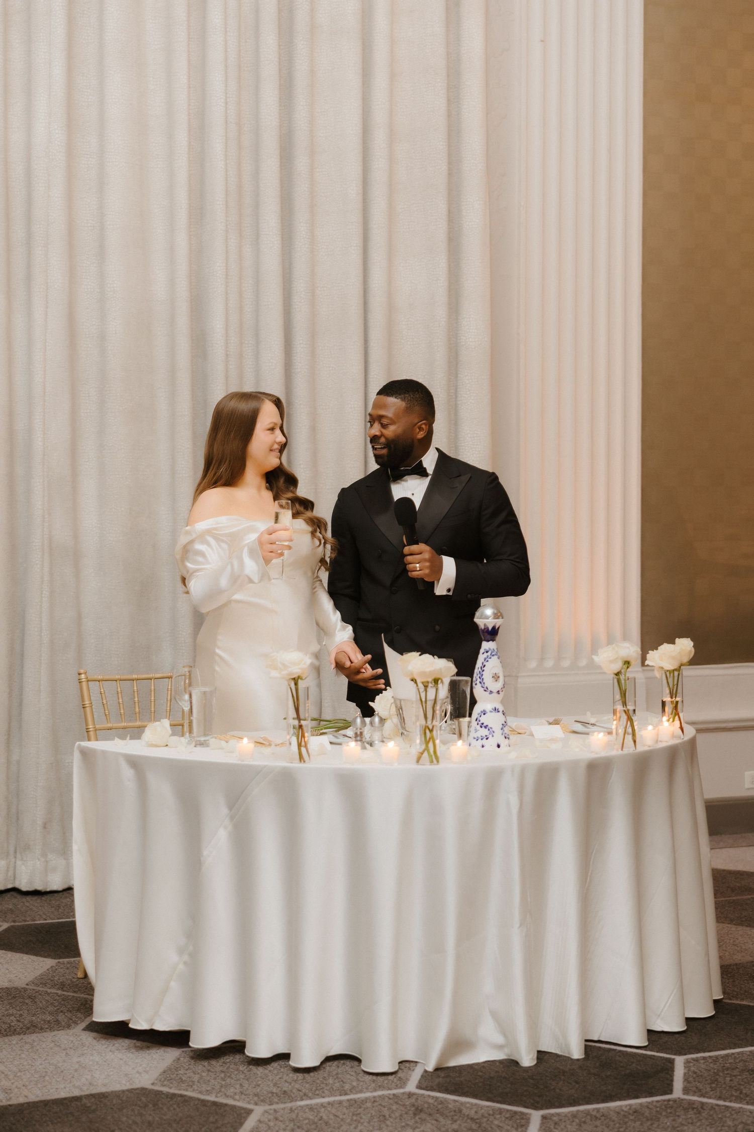 Bride and groom stand at elegant white sweetheart table with floral decor during wedding reception.