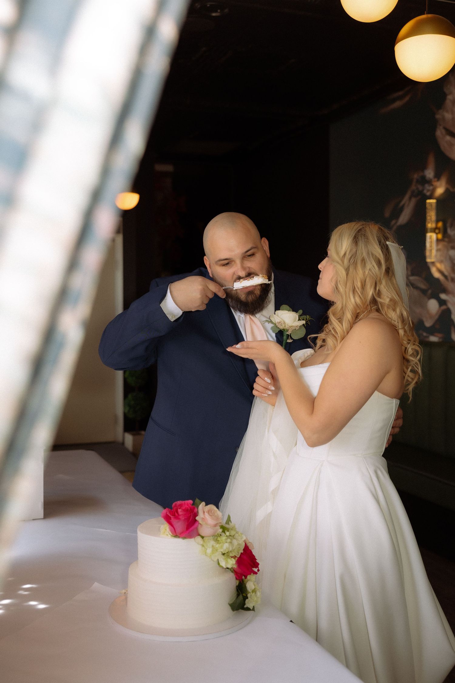Bride and groom sharing a joyful cake tasting moment at their wedding reception beside a white tiered cake.