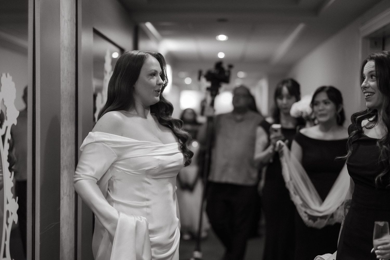 Bride in elegant white off-shoulder gown standing in hallway during wedding reception in black and white photo.