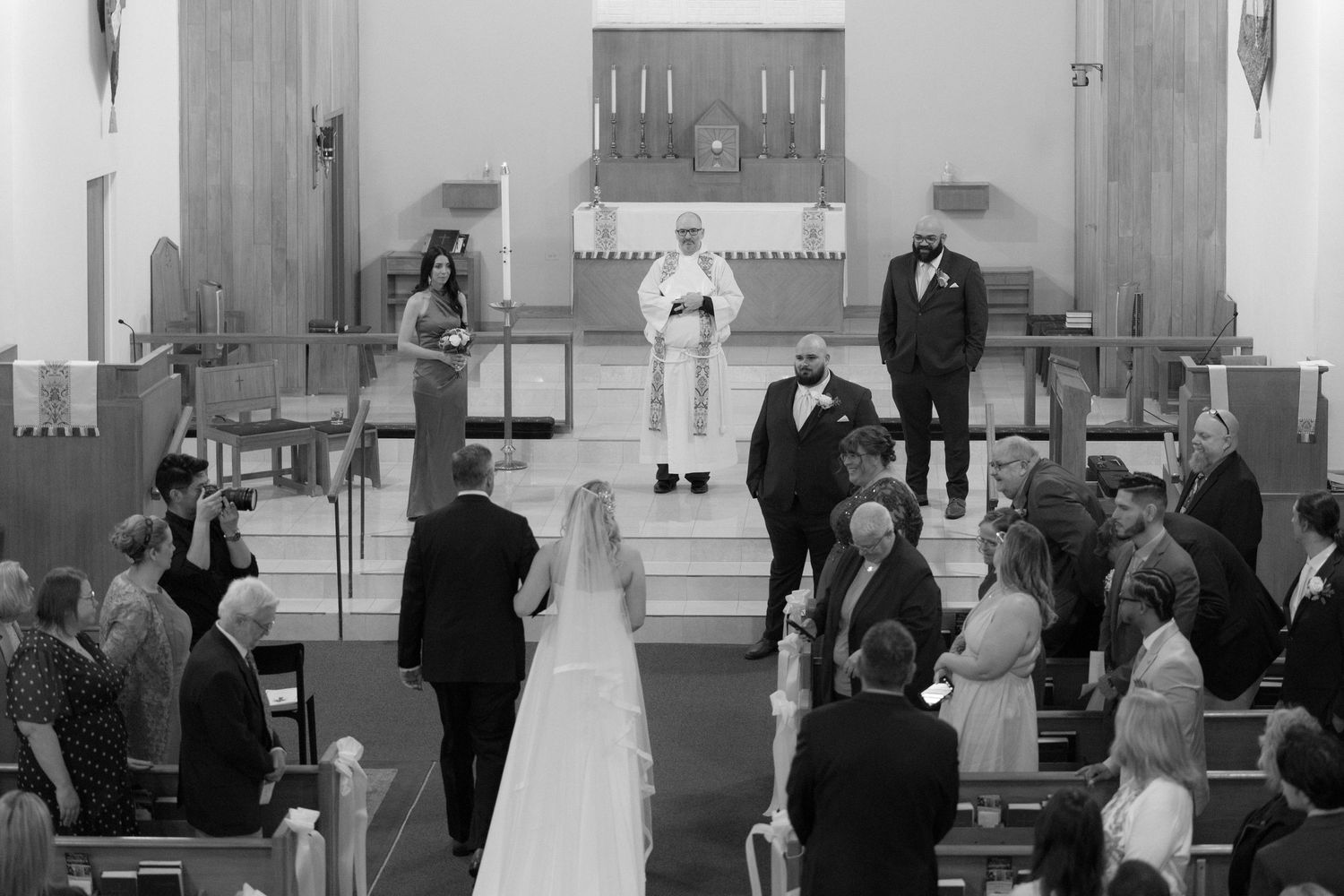 Black and white wedding ceremony photo inside a church with guests seated in pews and officiant at the altar.