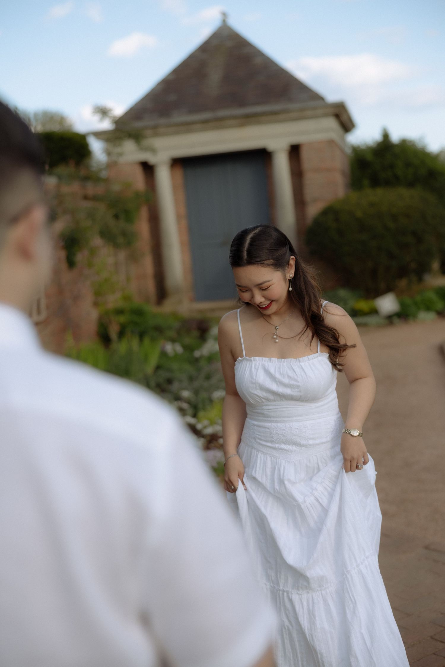 Woman in white sundress smiling in garden with historic stone pavilion backdrop.