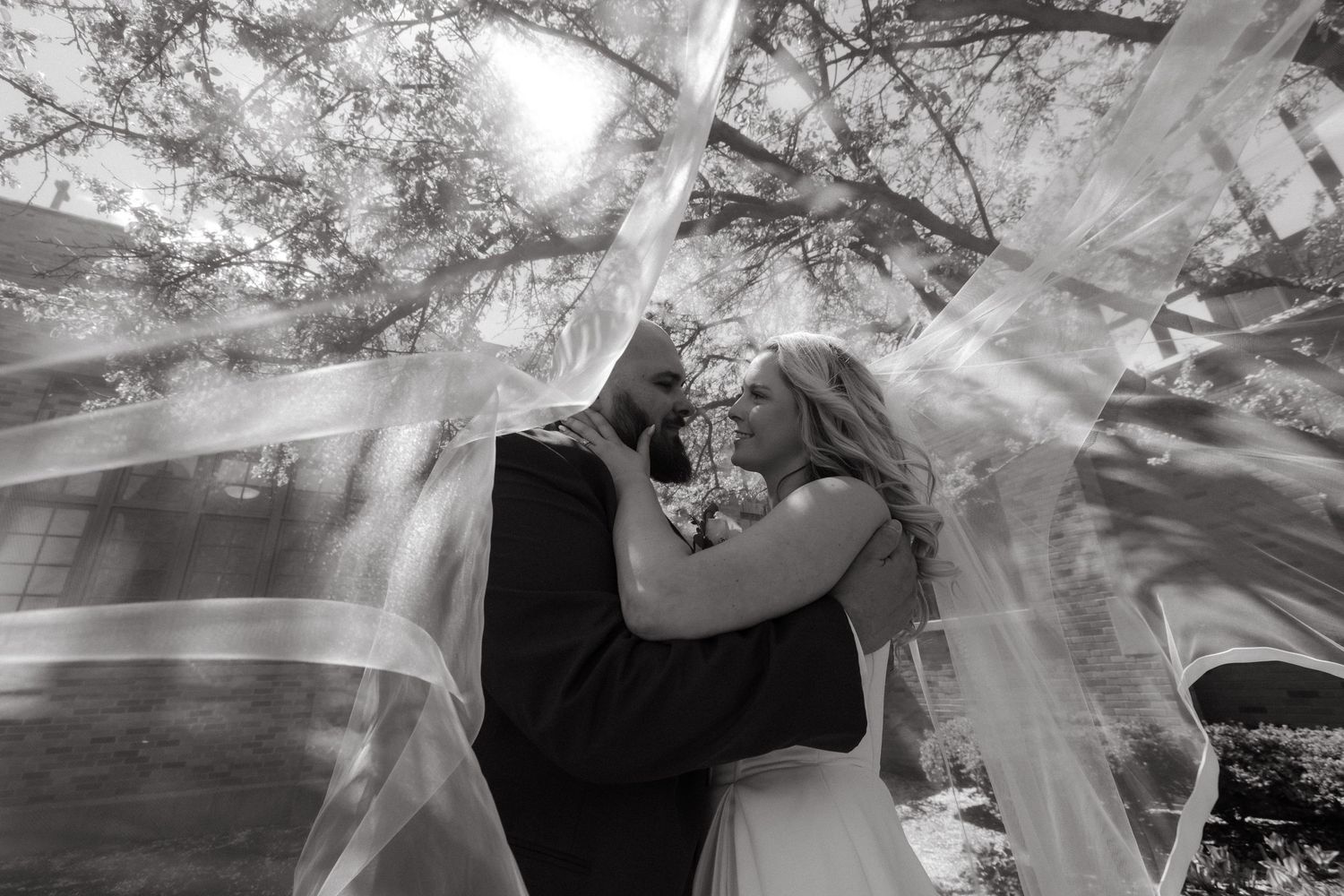 Black and white wedding photo of bride and groom kissing under flowing veil outdoors.