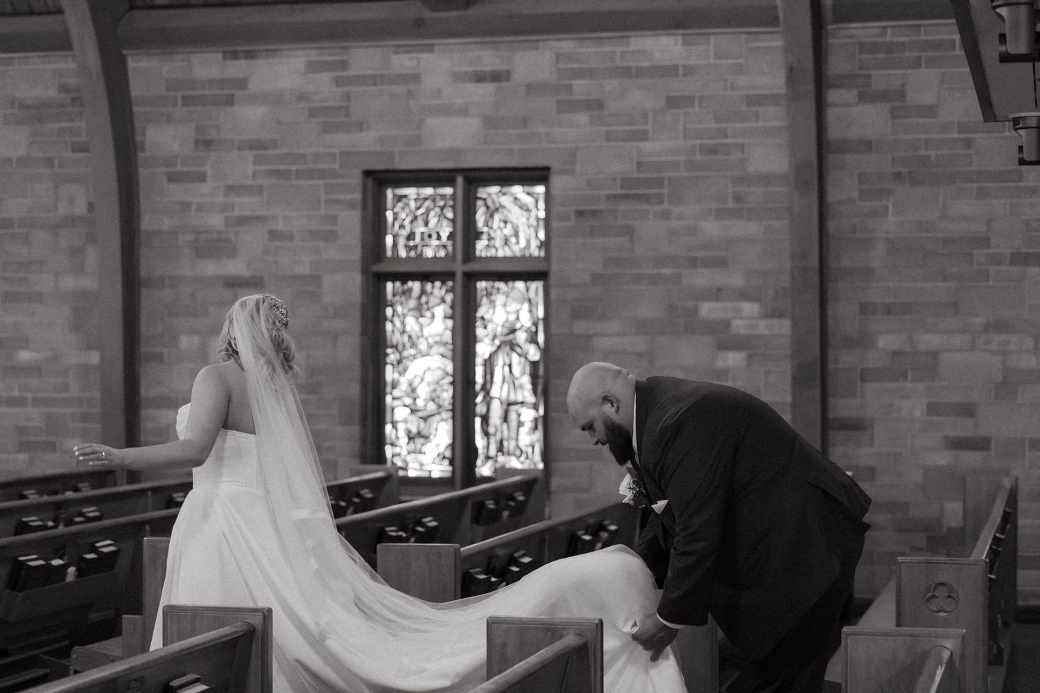 Black and white photo of a bride in veil with an older man bowing in a church pew during a wedding ceremony.