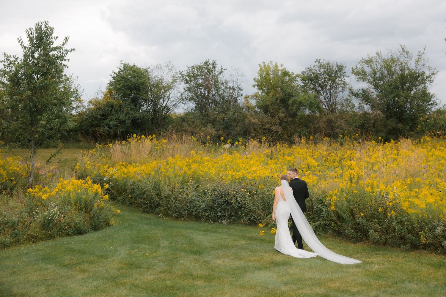 Bride and groom walking through a lush green field bordered by vibrant yellow wildflowers under a cloudy sky.