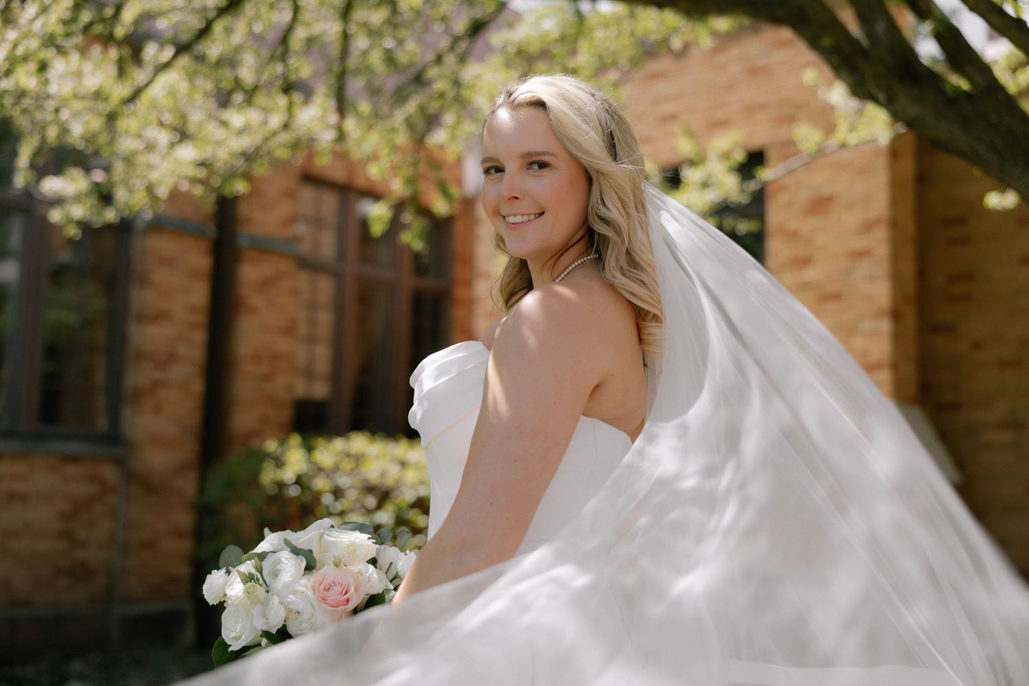 Smiling bride in white strapless gown and veil holds bouquet of pink and white flowers outdoors near brick building.