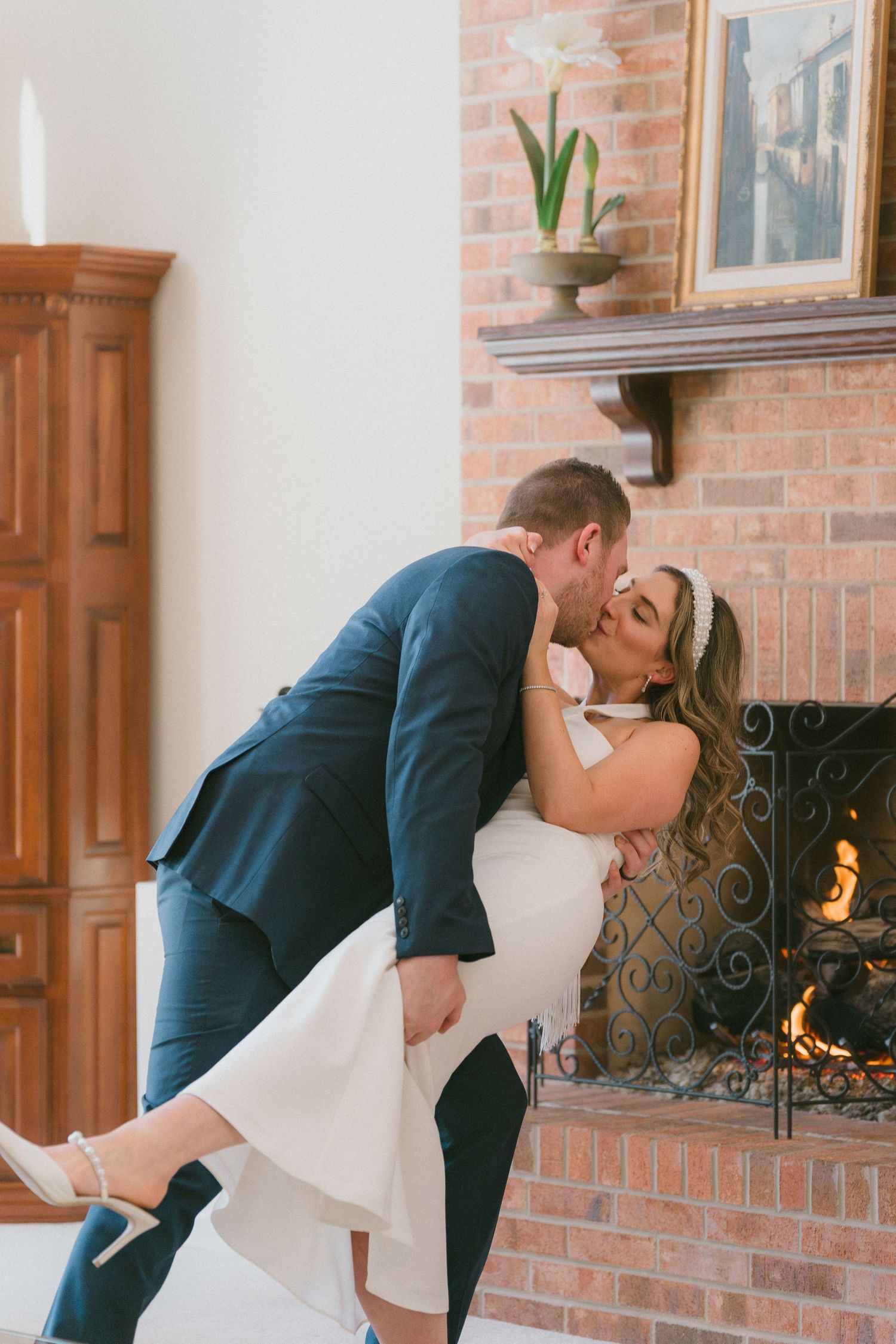 Groom dipping bride for a romantic kiss in front of a cozy brick fireplace at their wedding.